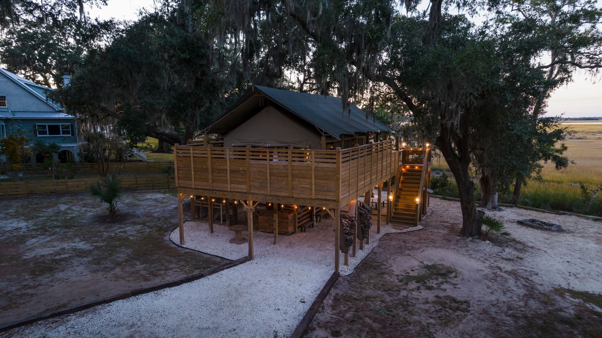 Wooden elevated cabin with a deck, lit by string lights, in a field with trees.