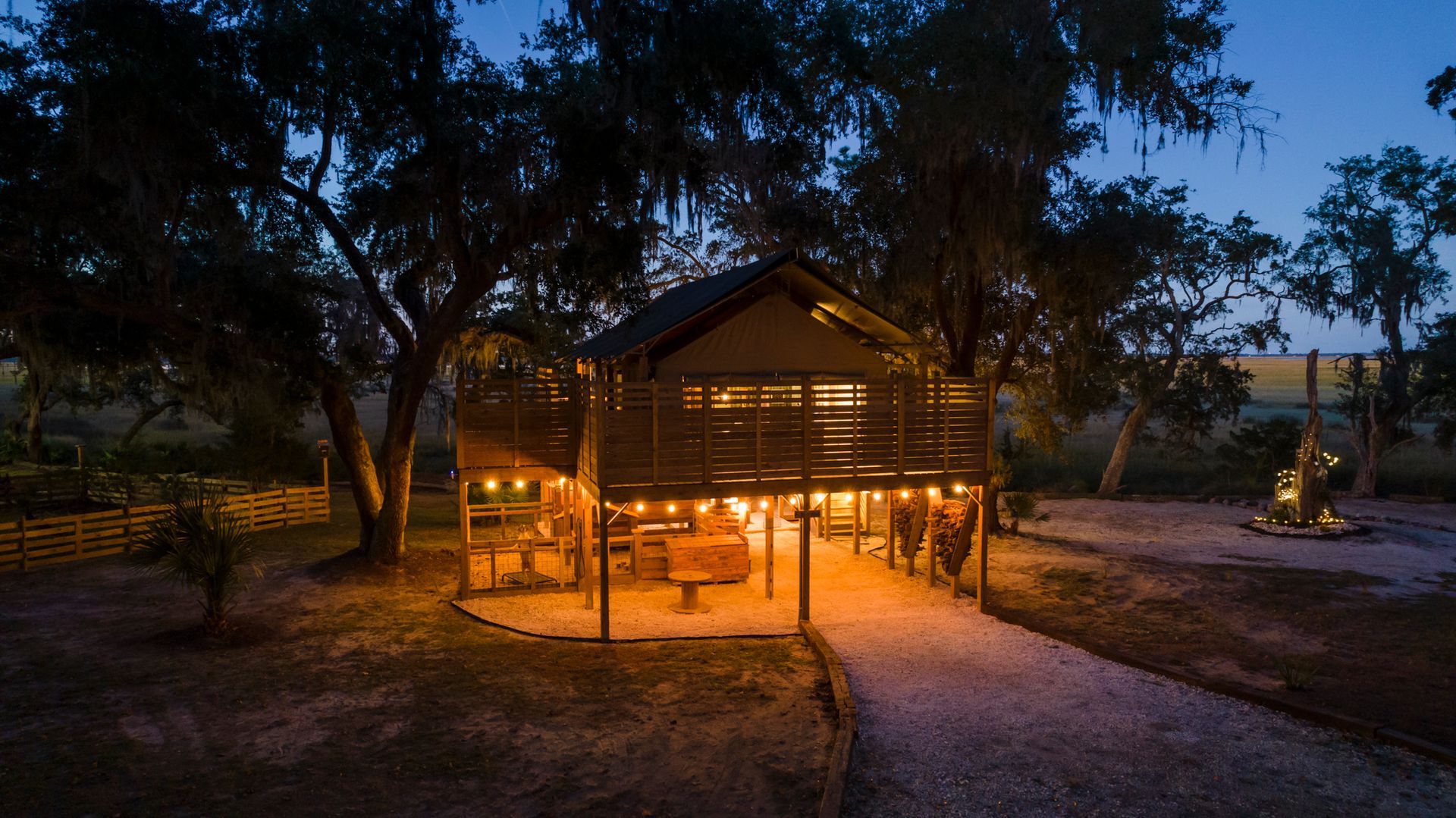 Lit-up wooden structure on stilts with string lights, gravel path, and surrounding trees at dusk.