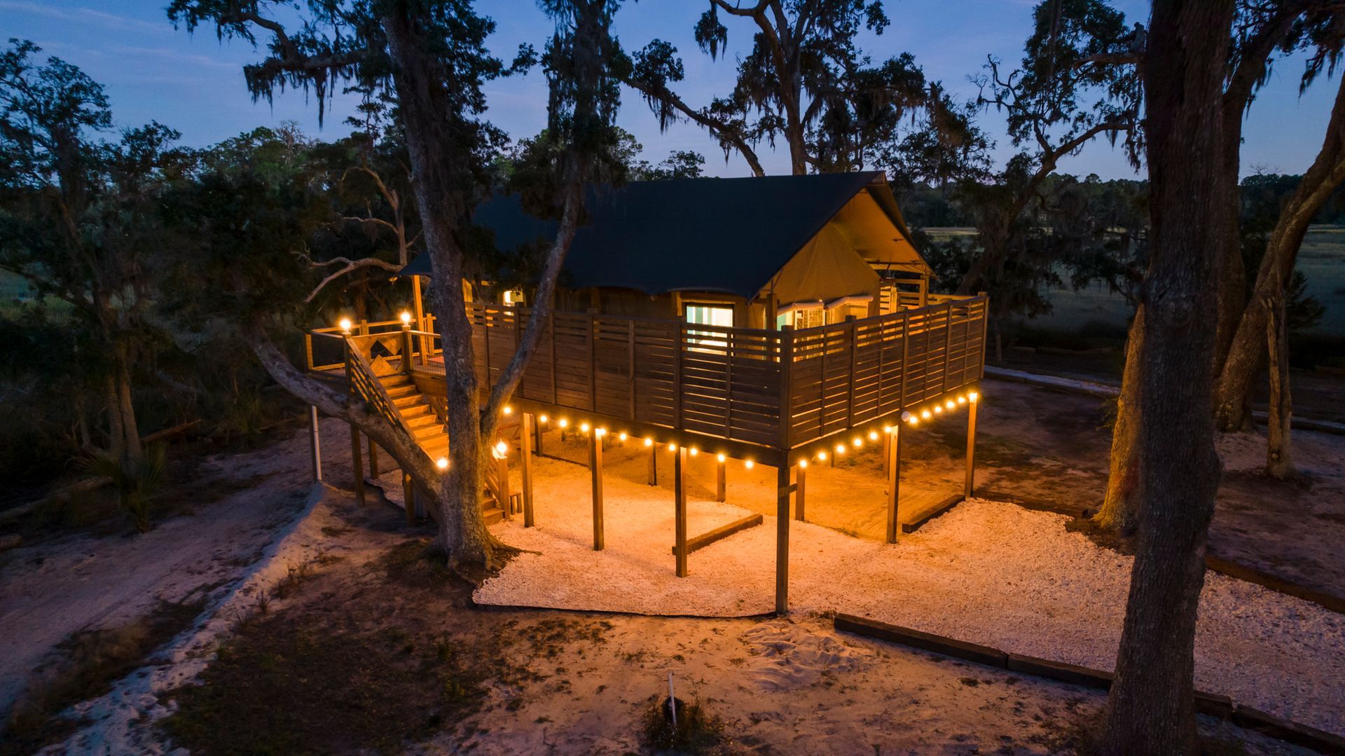 Elevated cabin lit with string lights at dusk; steps lead up, surrounded by trees.