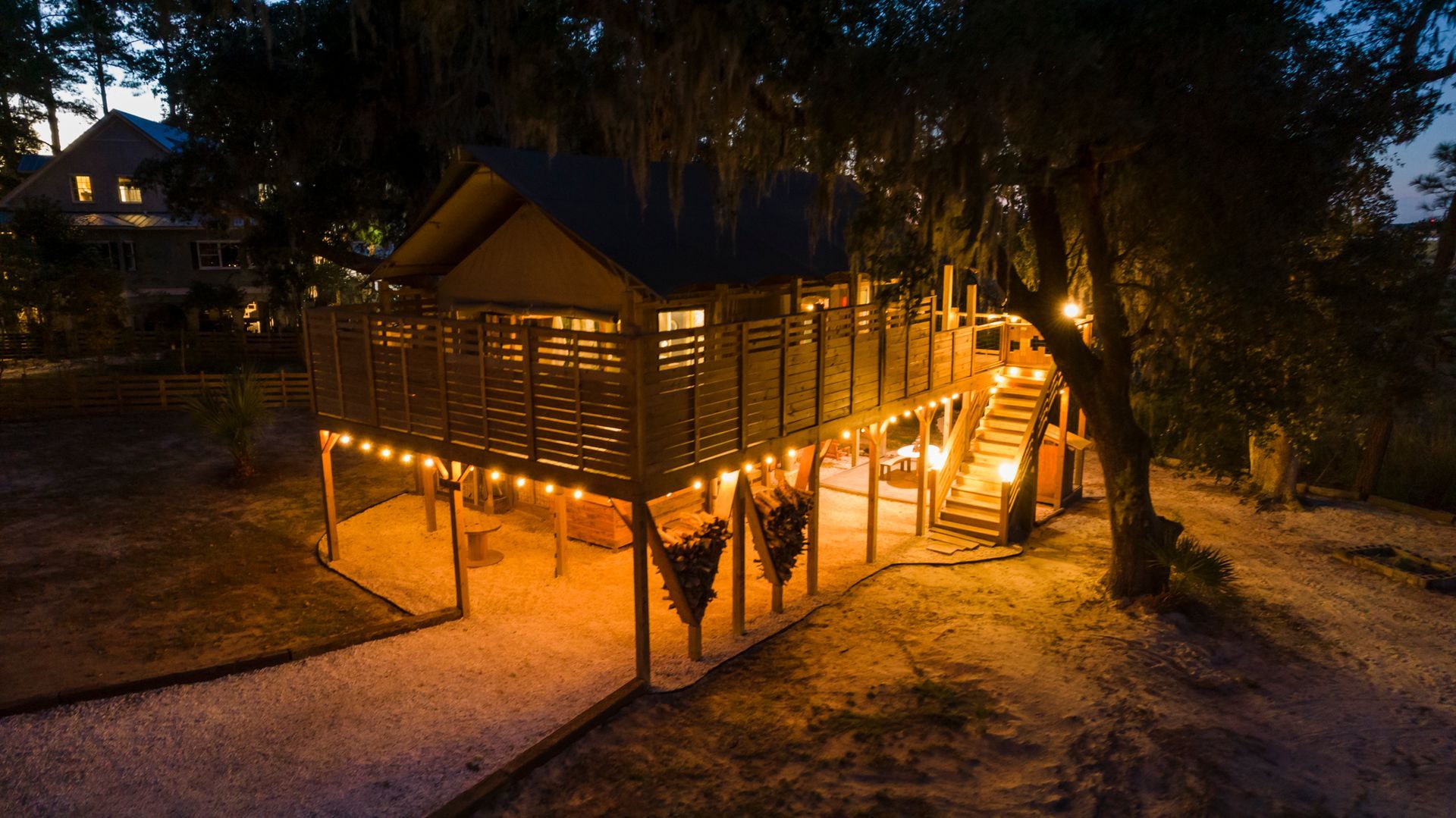 Lit-up elevated wooden structure at night; string lights, stairs, and a gravel path are visible.
