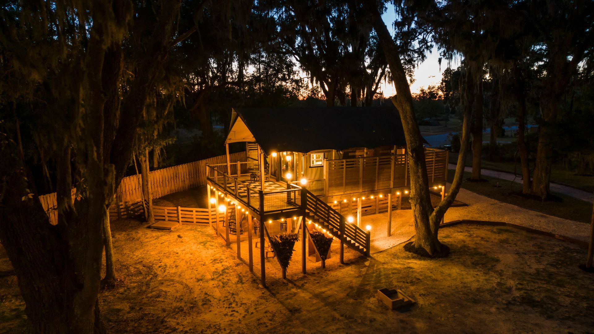 Wooden cabin with string lights on a raised deck, surrounded by trees at dusk.