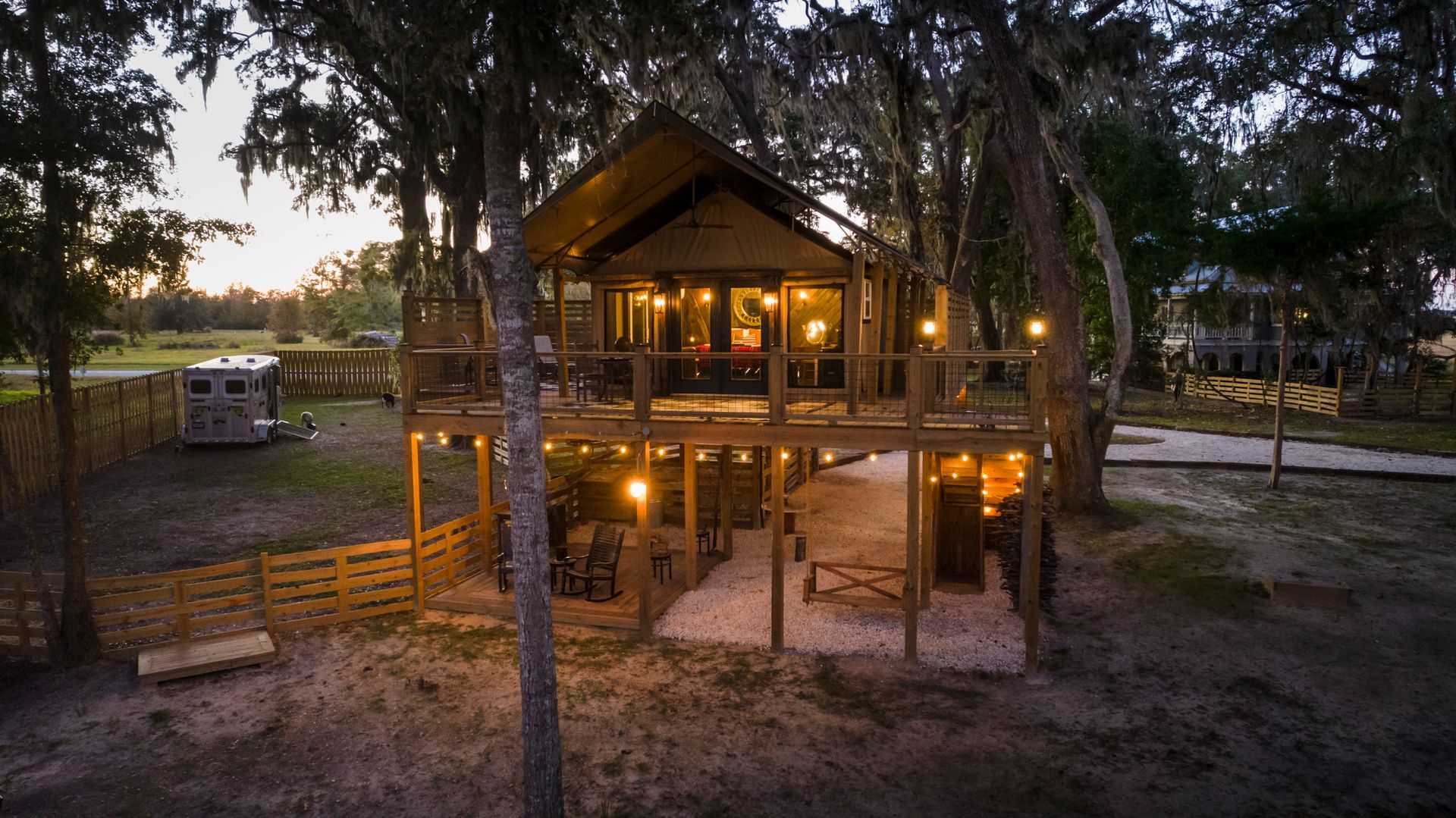 Treehouse with wooden deck, string lights, and camper in a wooded area.