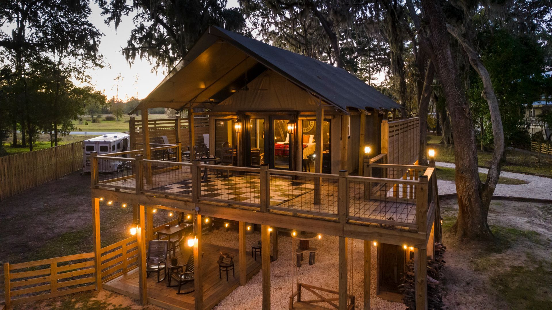 Wooden cabin with two-story deck, string lights, and a dark roof, in a wooded outdoor setting.