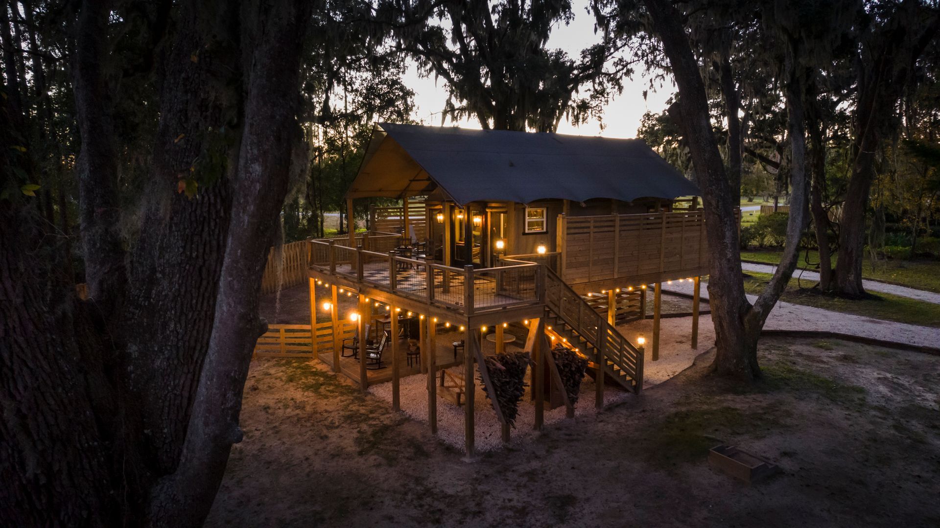 Elevated wooden treehouse with string lights, surrounded by trees and a sandy ground; dusk setting.