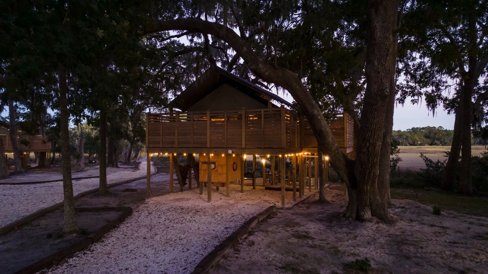 Treehouse lit with string lights at dusk; gravel path leads to structure.