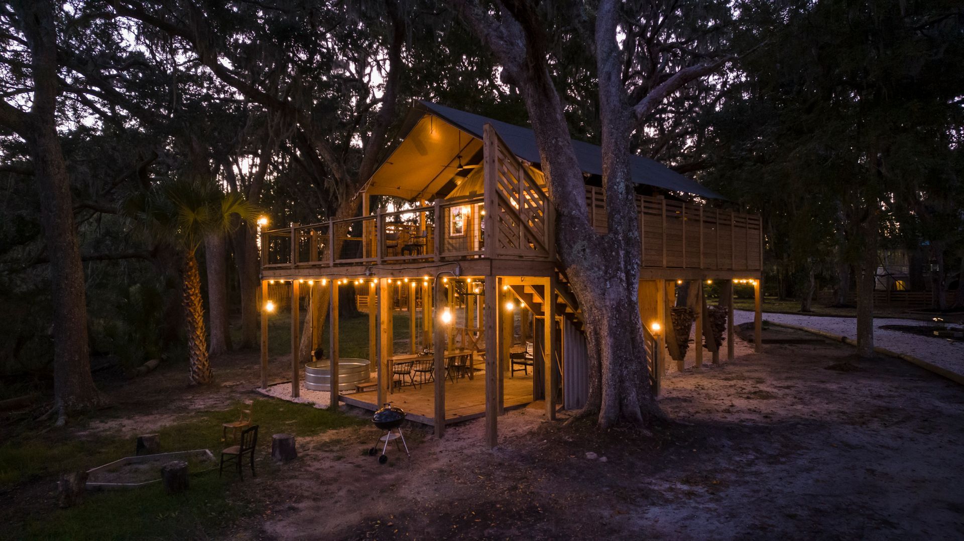 Treehouse with string lights illuminated at dusk. Wood and canvas structure built around a large tree, surrounded by foliage.