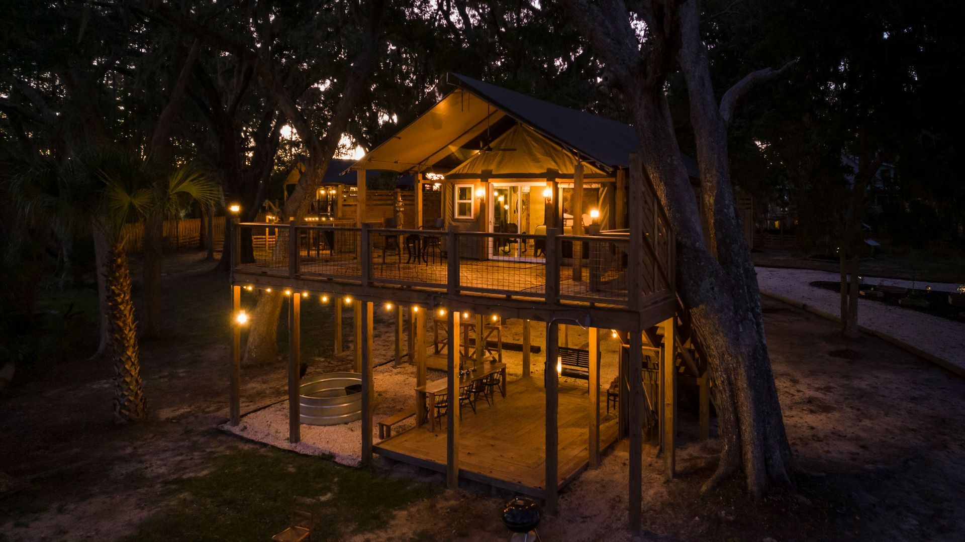Lit-up elevated wooden treehouse at night, surrounded by trees and string lights.