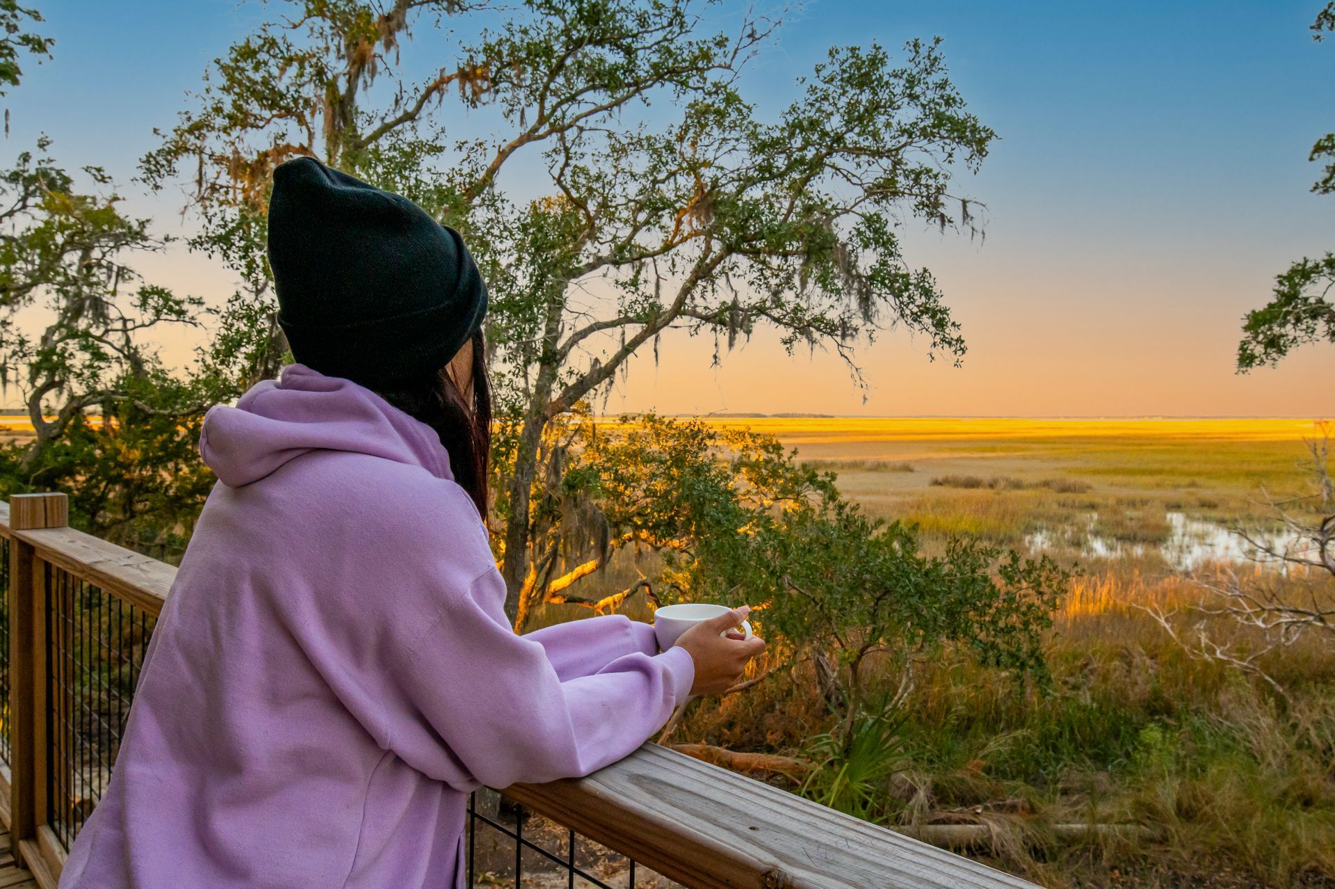 Person on a wooden deck looking at marshland at sunset, holding a mug.