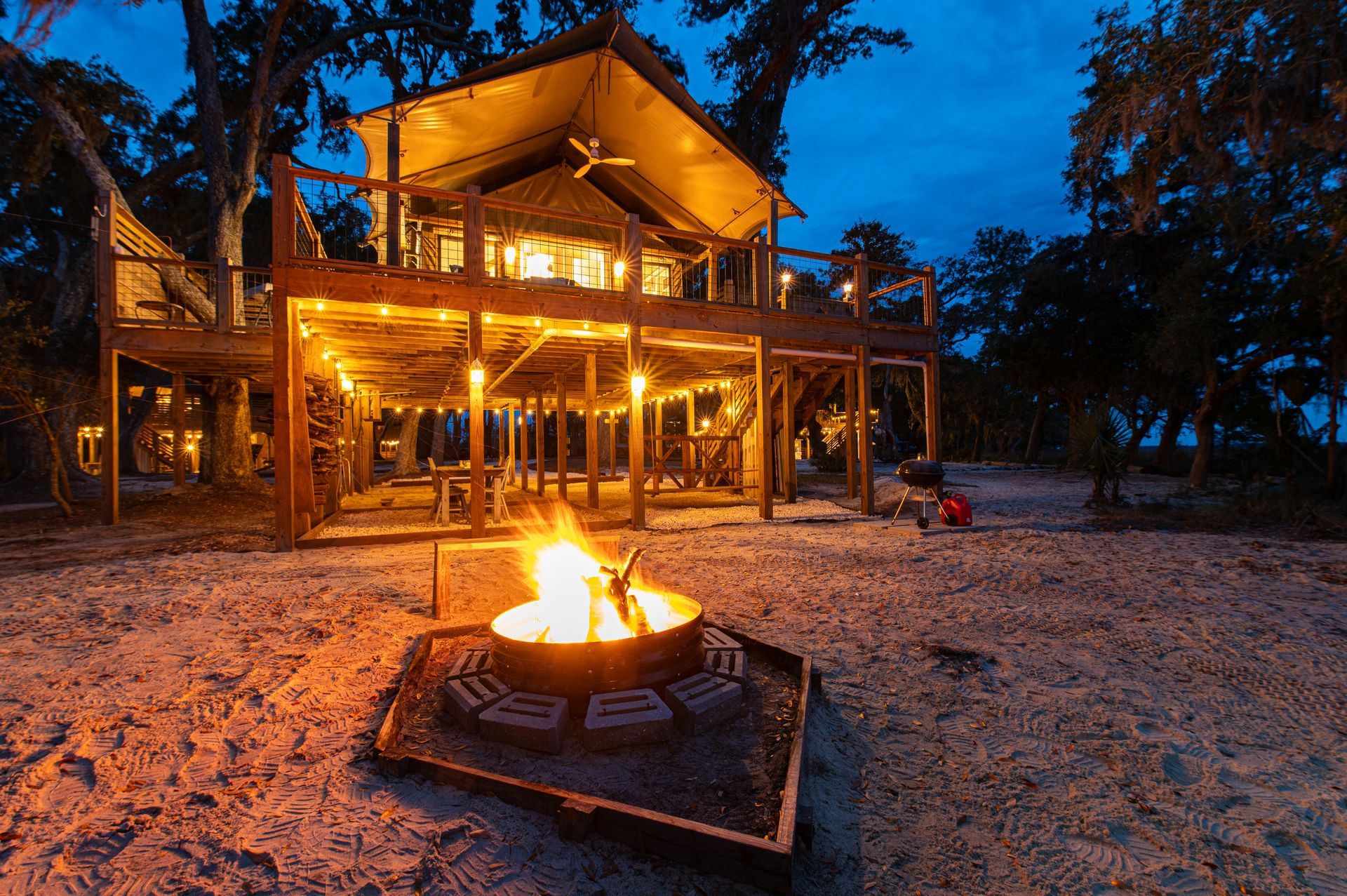 Lit cabin with fire pit on a sandy beach at dusk.