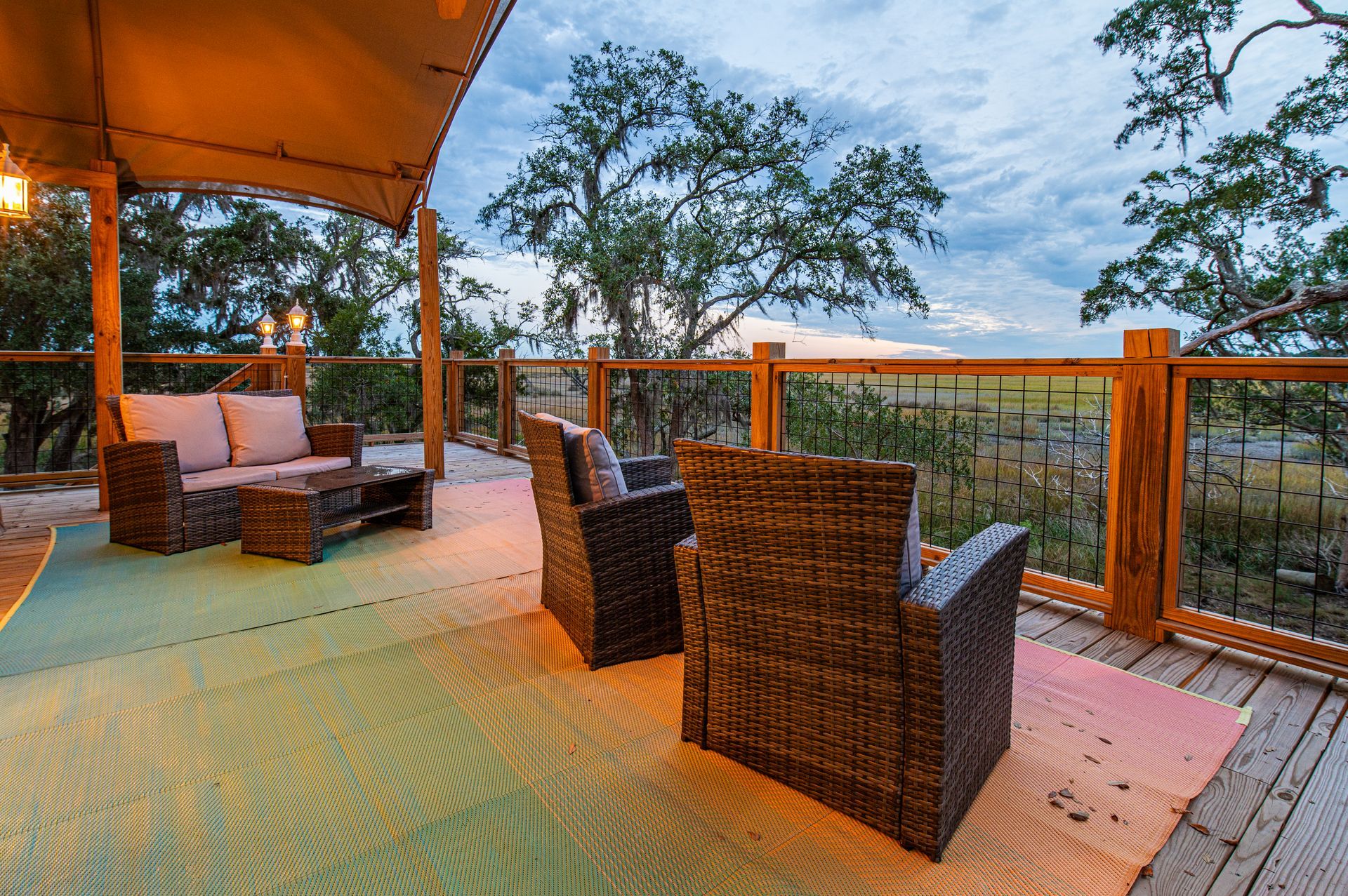 Outdoor deck with wicker furniture, rug, and view of trees under a cloudy sky.