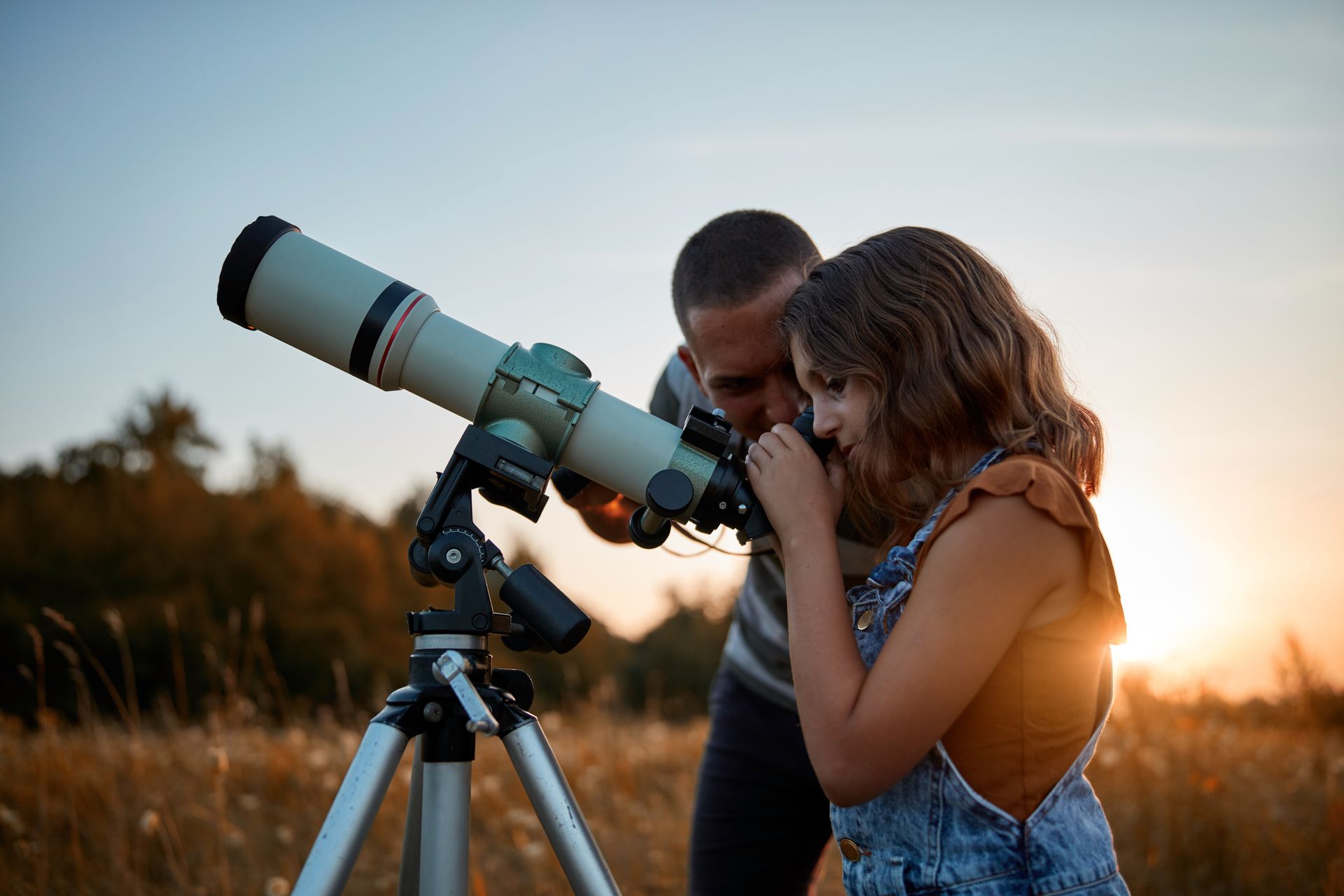 Man and child looking through a telescope in a field at sunset.