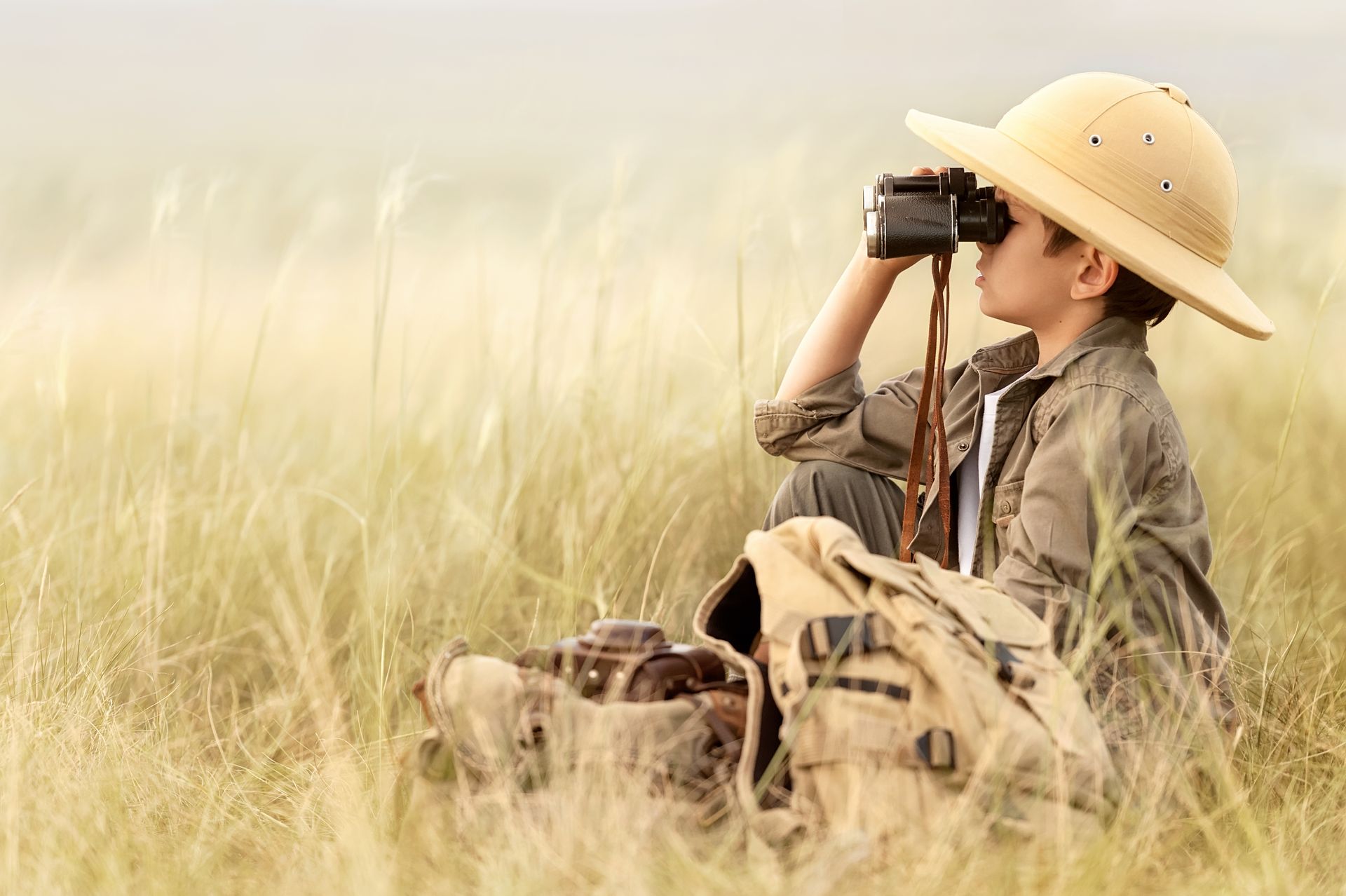 A young boy is sitting in a field looking through binoculars
