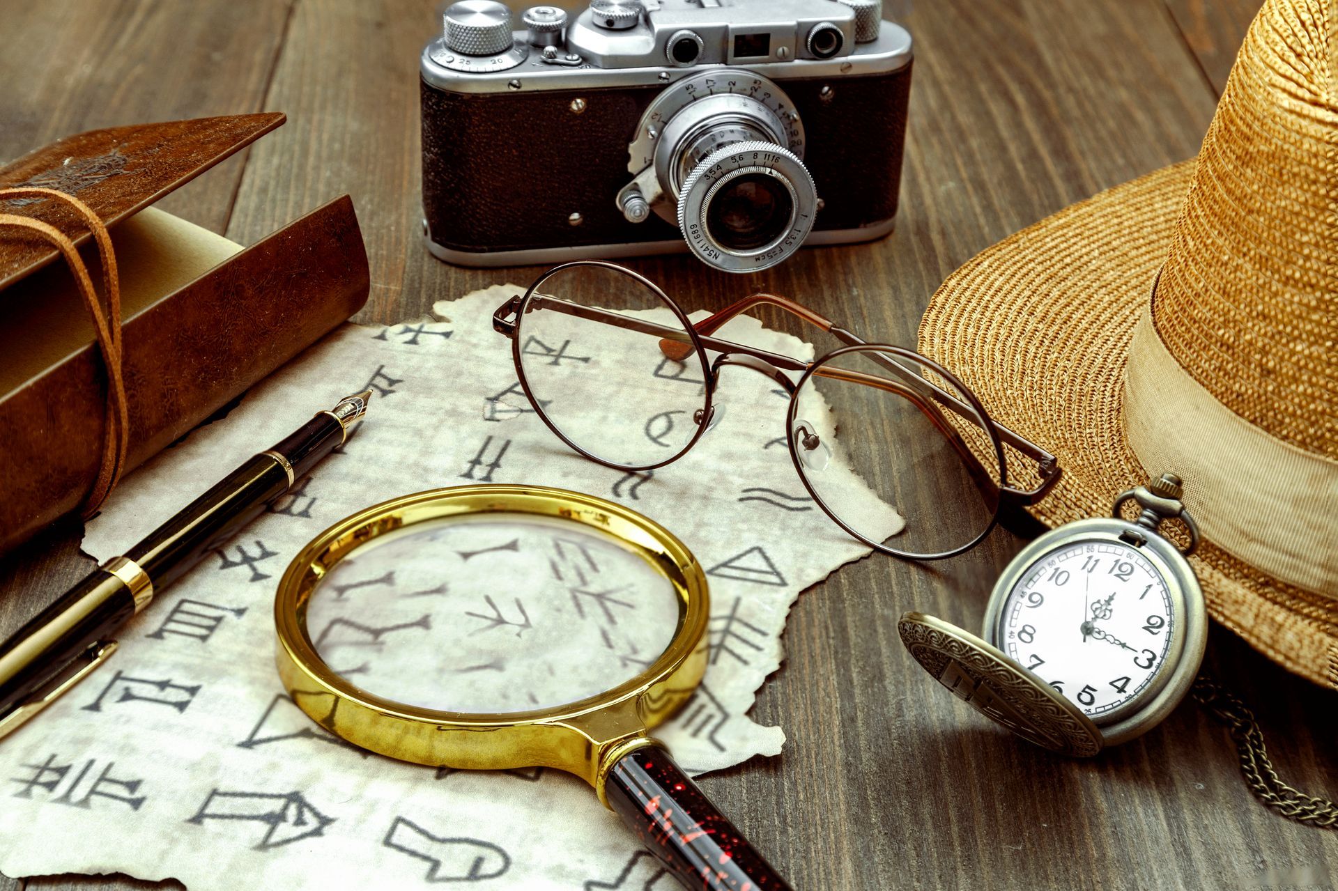 A camera , magnifying glass , glasses , a pocket watch , and a hat are on a wooden table.