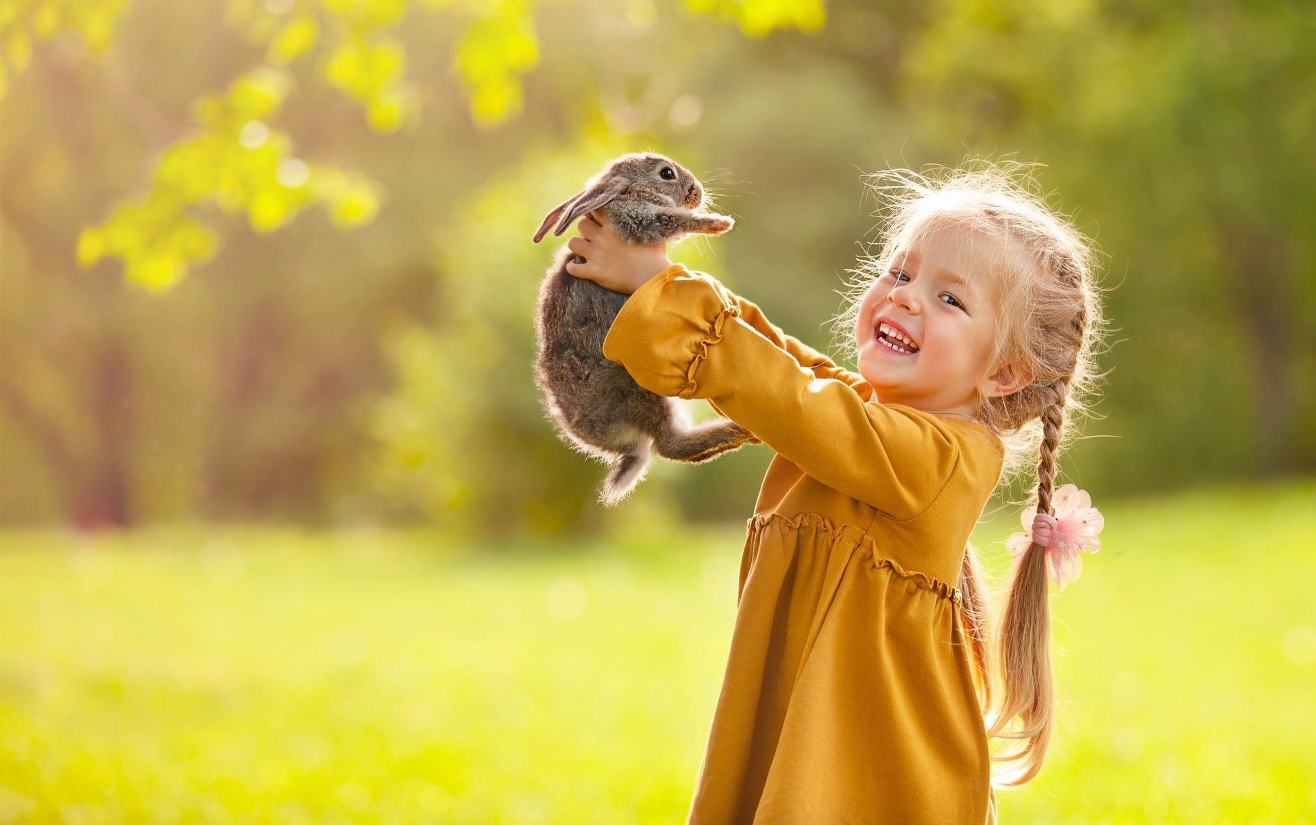 Young girl in mustard dress holding a bunny outdoors, laughing. Bright green field and trees in the background.