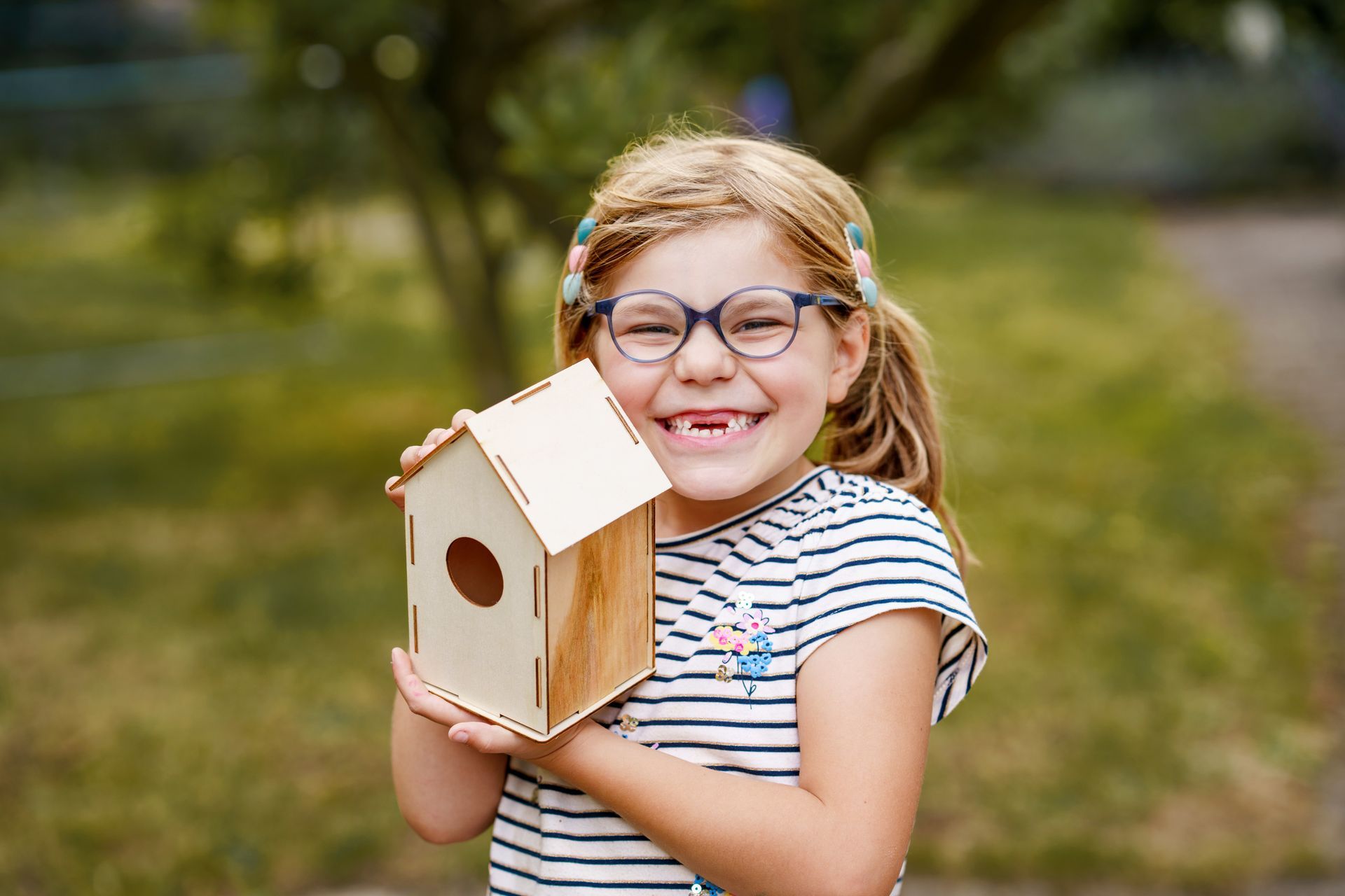 Girl with glasses smiling, holding a wooden birdhouse. Outdoors, blurred green background.