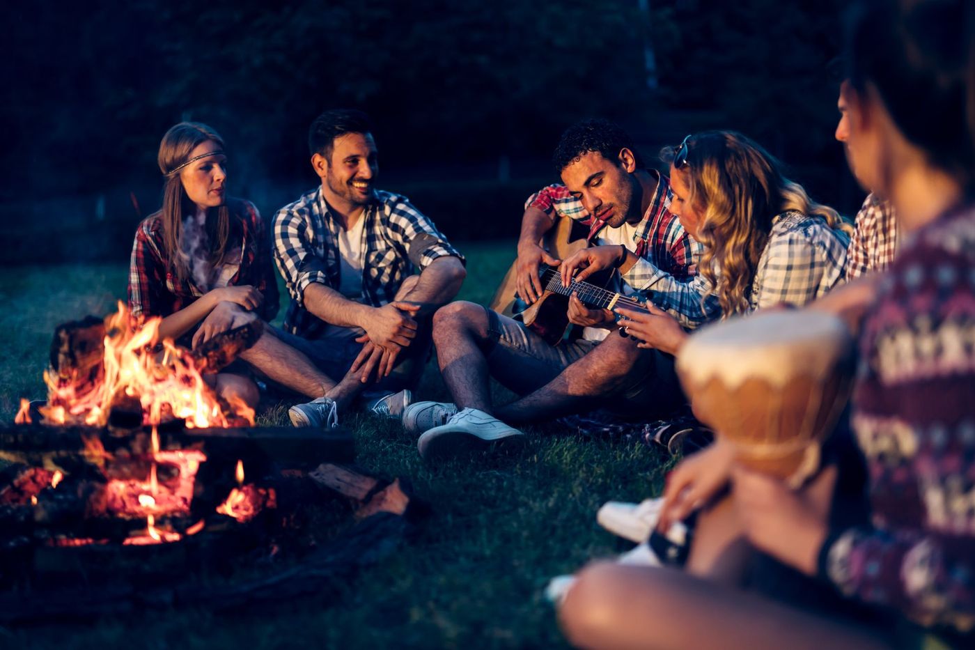 Group of people around a campfire at night, with one playing a guitar and another holding a drum.