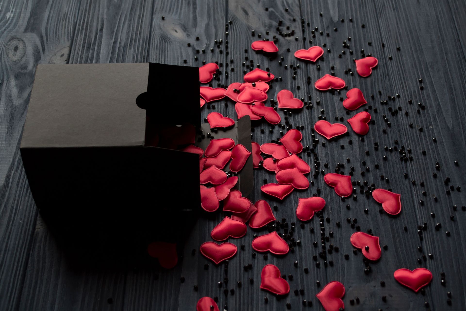 Red hearts are falling out of a black box on a wooden table.