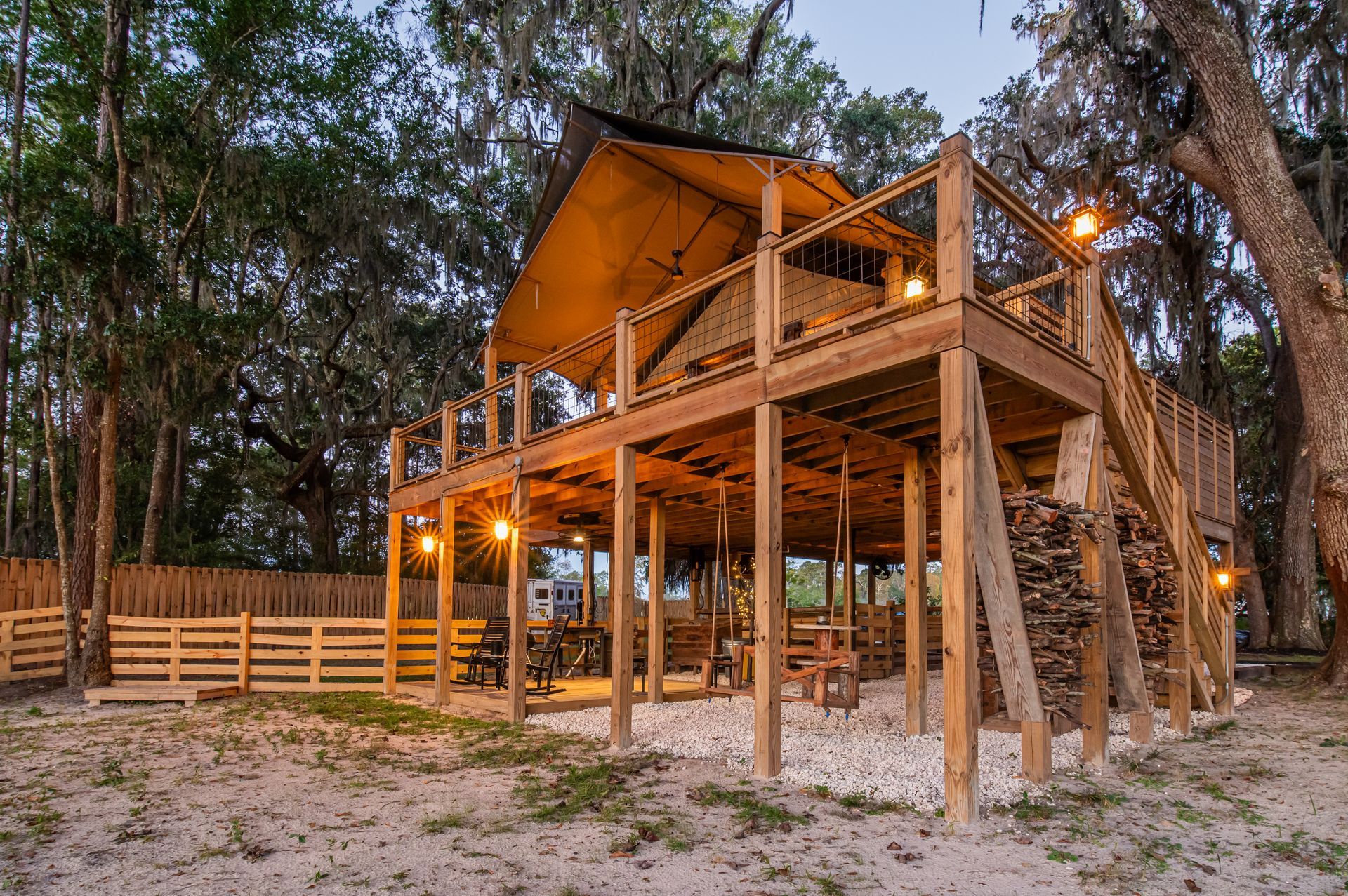 Wooden treehouse with a canvas roof, elevated above a sandy area, lit by string lights.