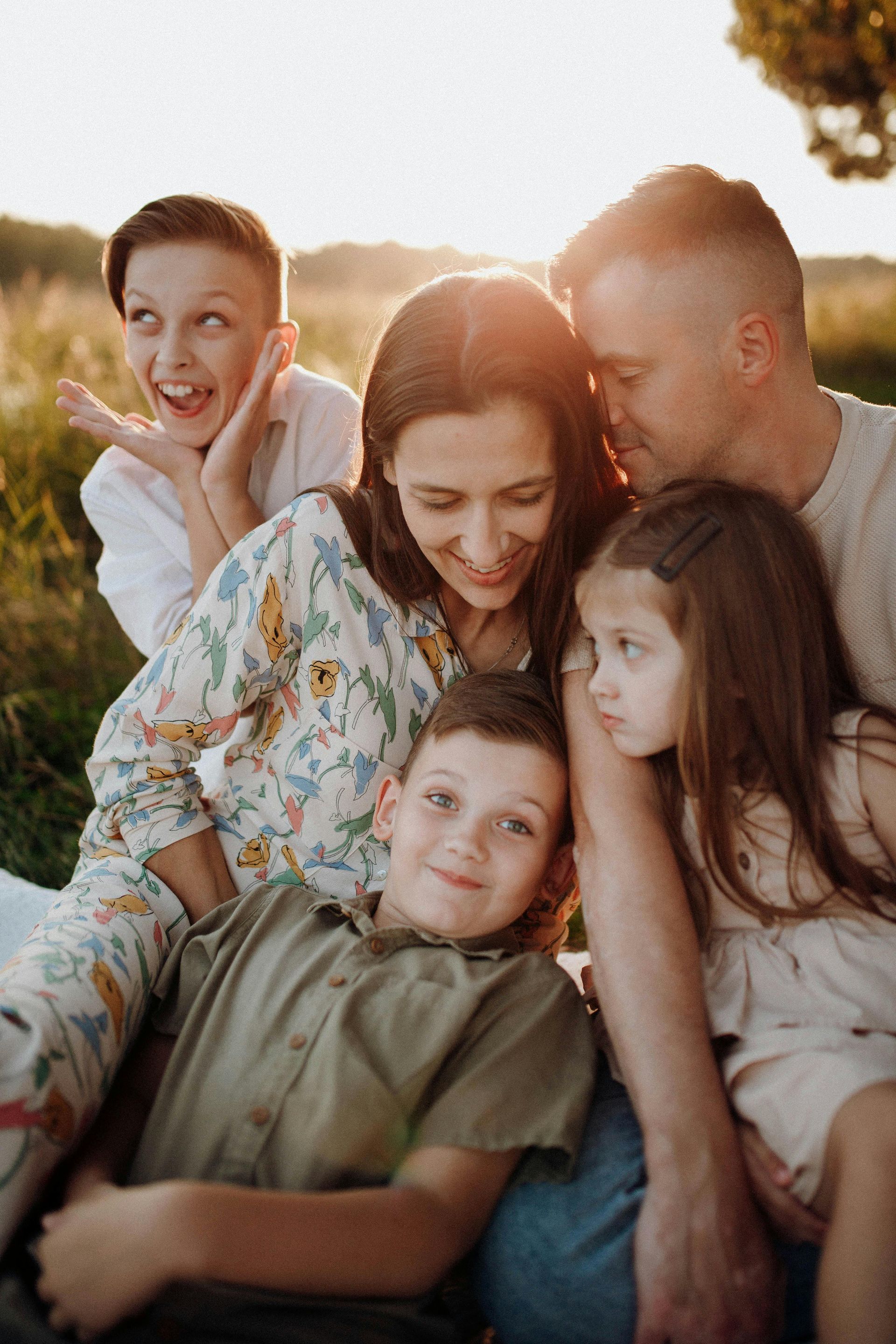 Family of five sitting outdoors in grass, smiling and looking down. Sun setting.
