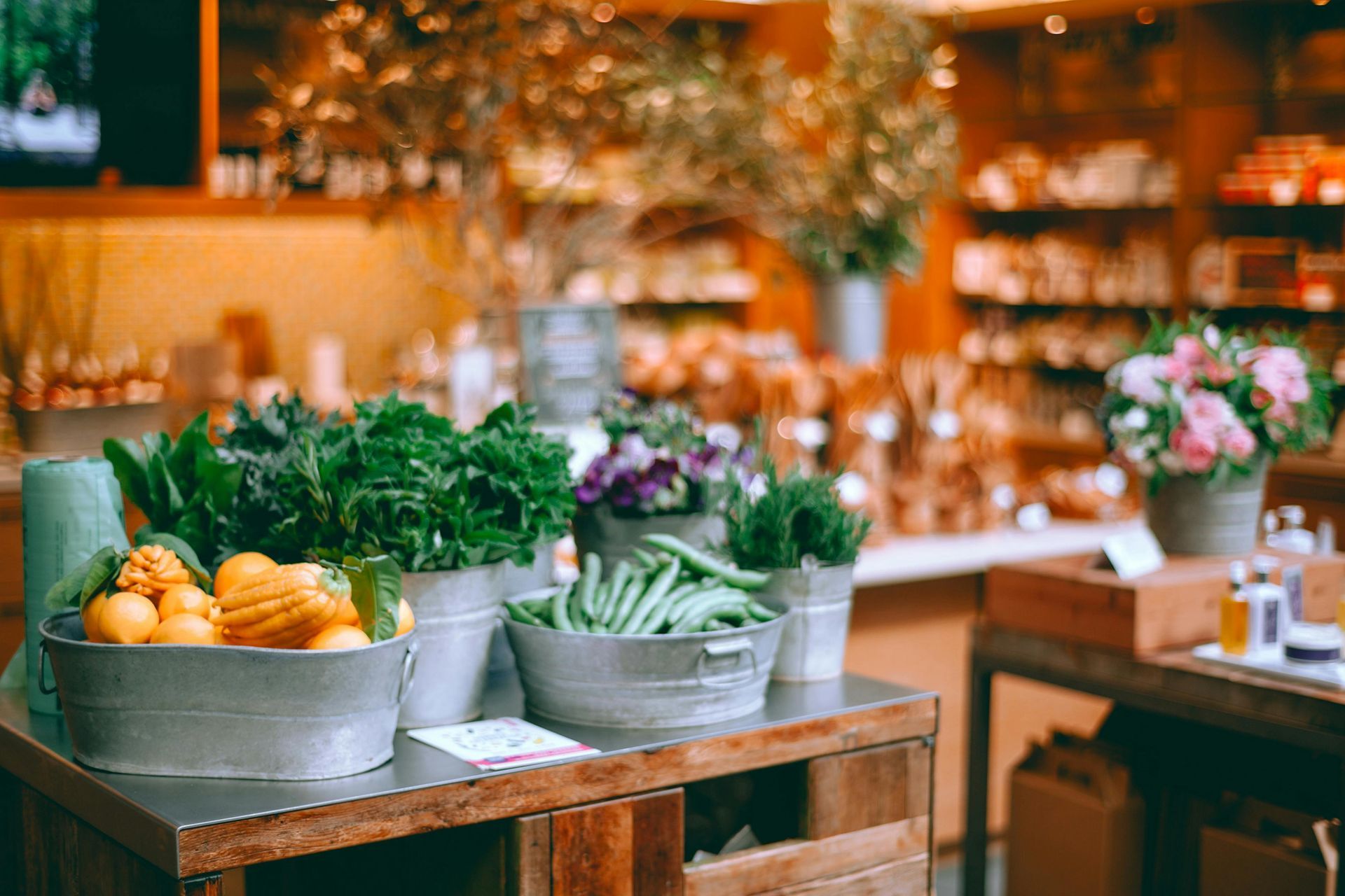 Produce stand with fresh produce in metal buckets, store interior with flowers, shelves, and soft lighting.