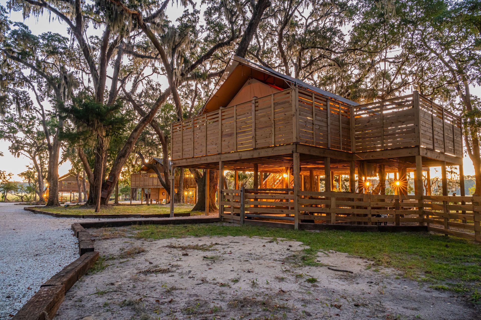 Wooden treehouse structure at sunset with surrounding trees and gravel pathway.