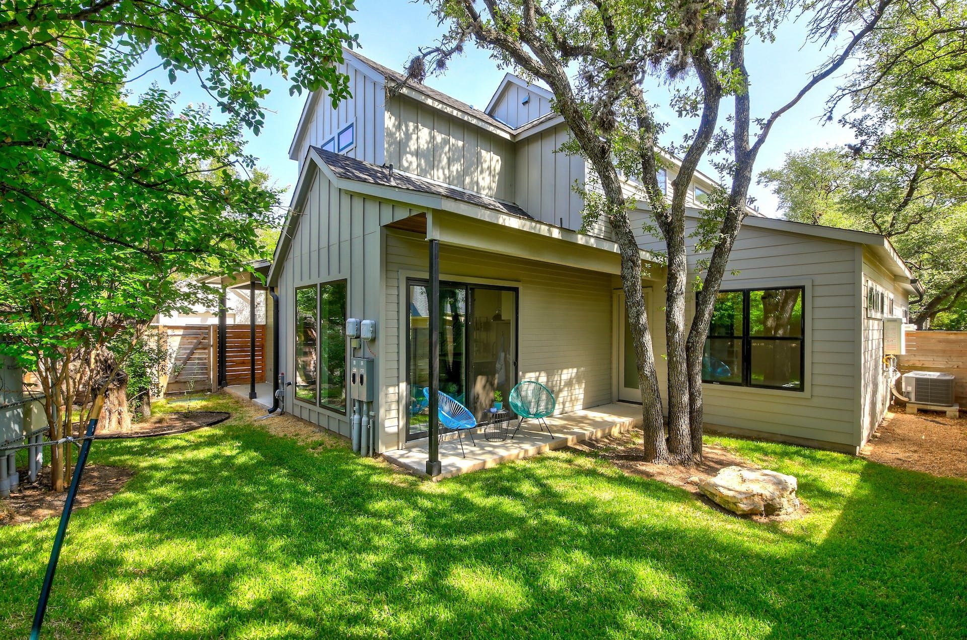 Backyard view of a two-story beige house with a covered patio, green grass, and trees.