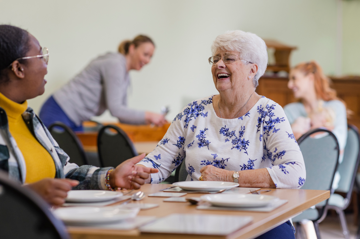 Two older women are sitting at a table with plates of food and laughing.