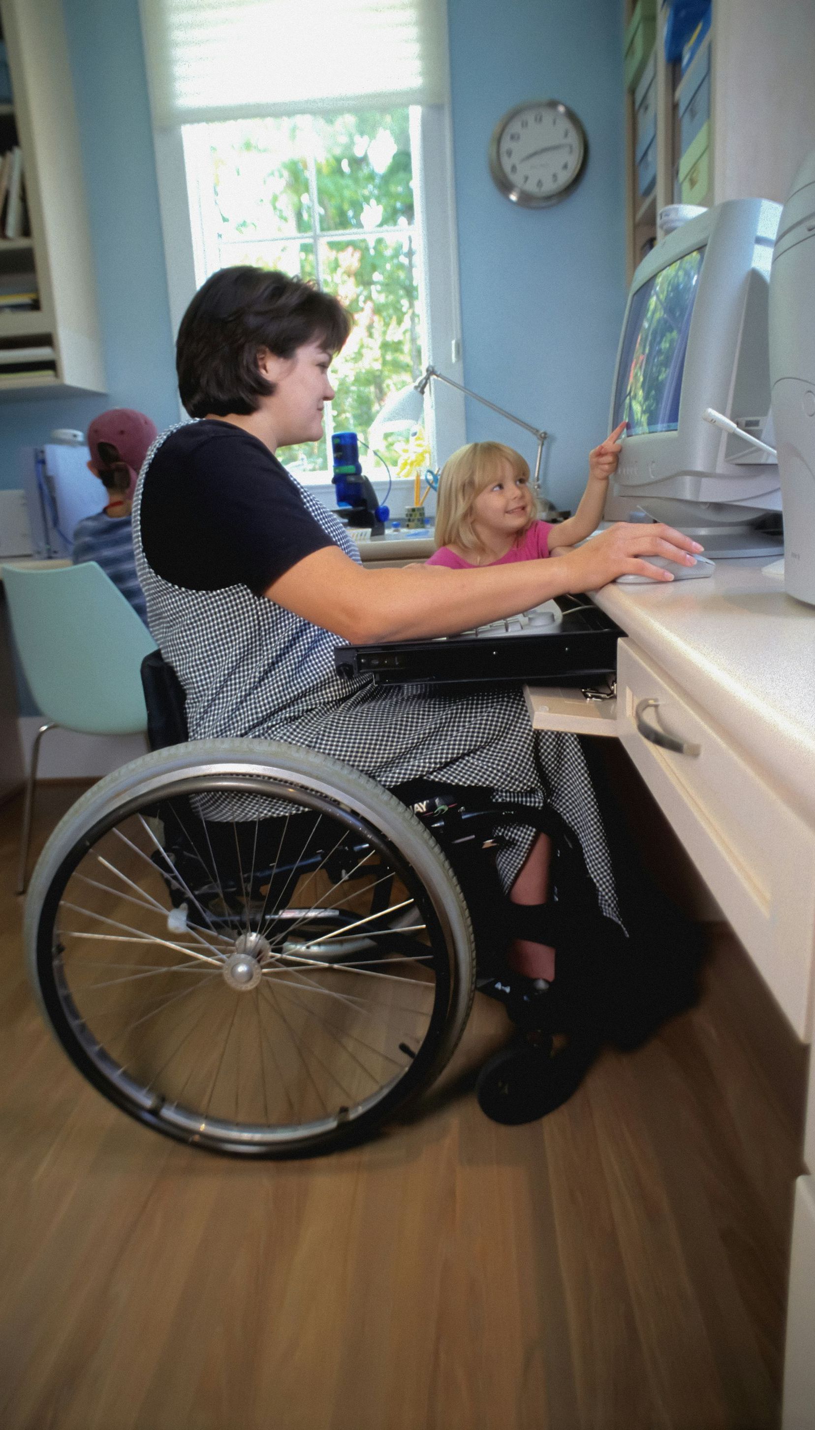 A woman in a wheelchair is using a computer while a little girl looks on.