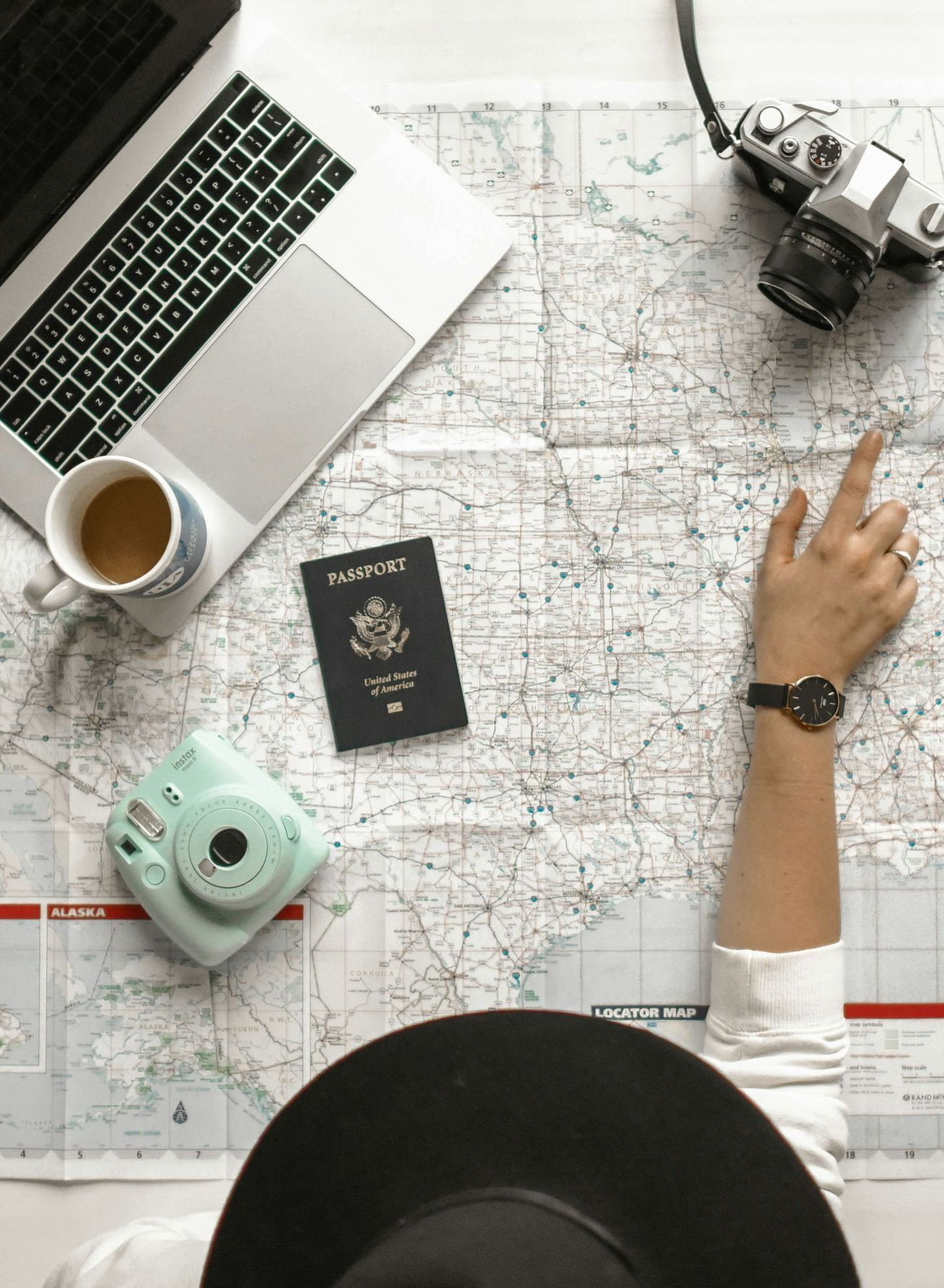 A person is sitting at a table with a map , passport , camera , and laptop.