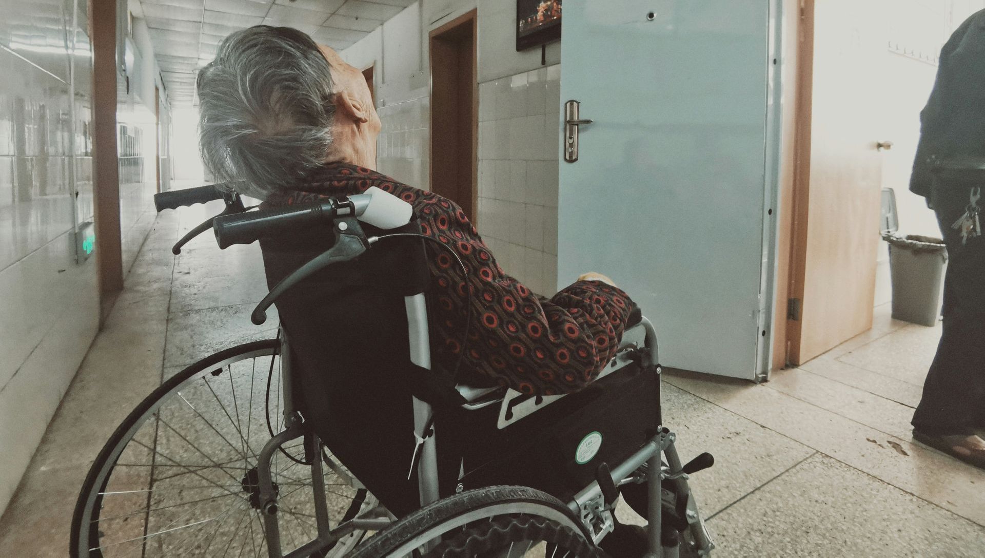An elderly woman is sitting in a wheelchair in a hospital hallway.