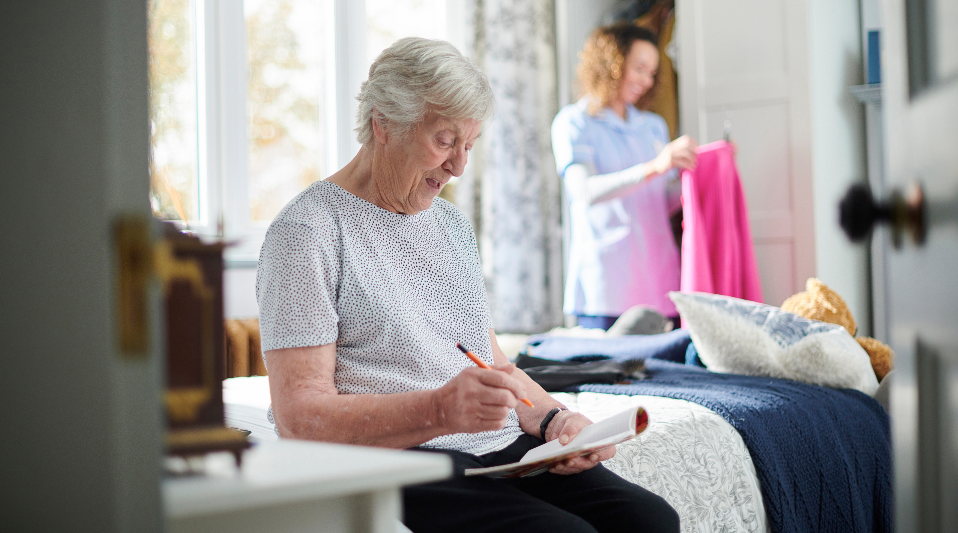 An elderly woman is sitting on a bed writing in a notebook.