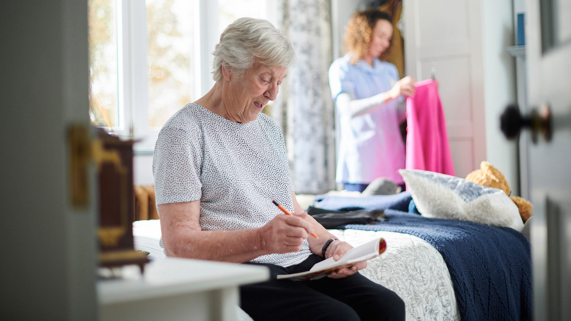 An elderly woman is sitting on a bed writing in a notebook.