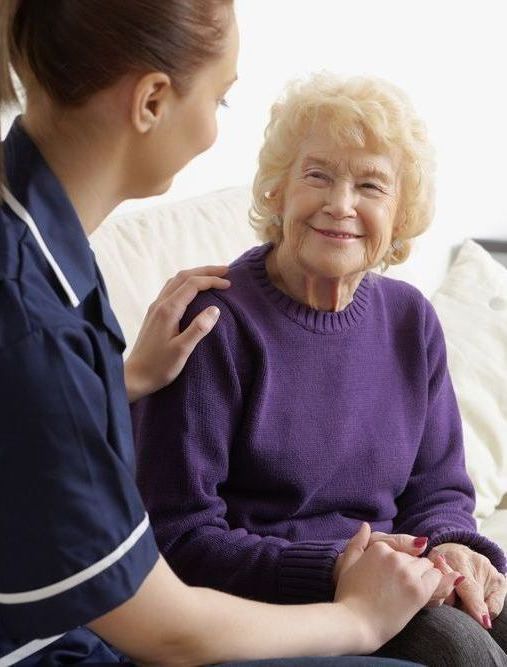 An elderly woman in a purple sweater is sitting on a couch with a nurse.