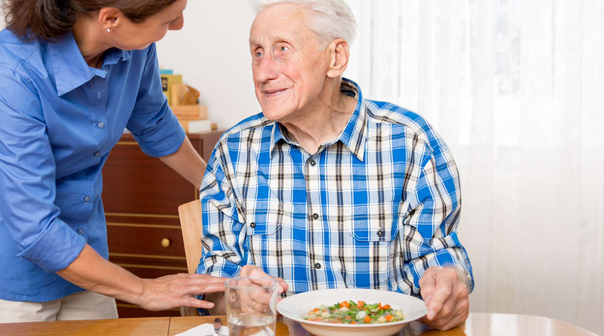 An elderly man is sitting at a table with a plate of food and a glass of water.