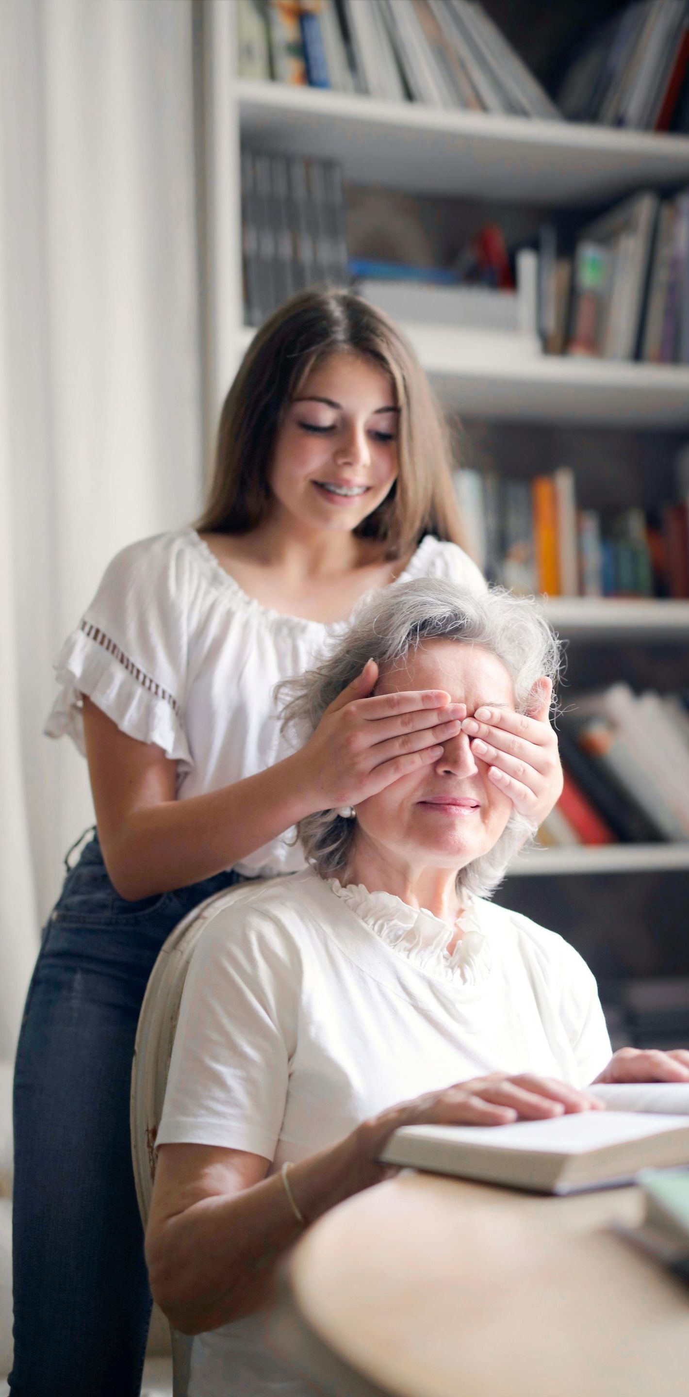 A young girl is covering an older woman 's eyes while she reads a book.