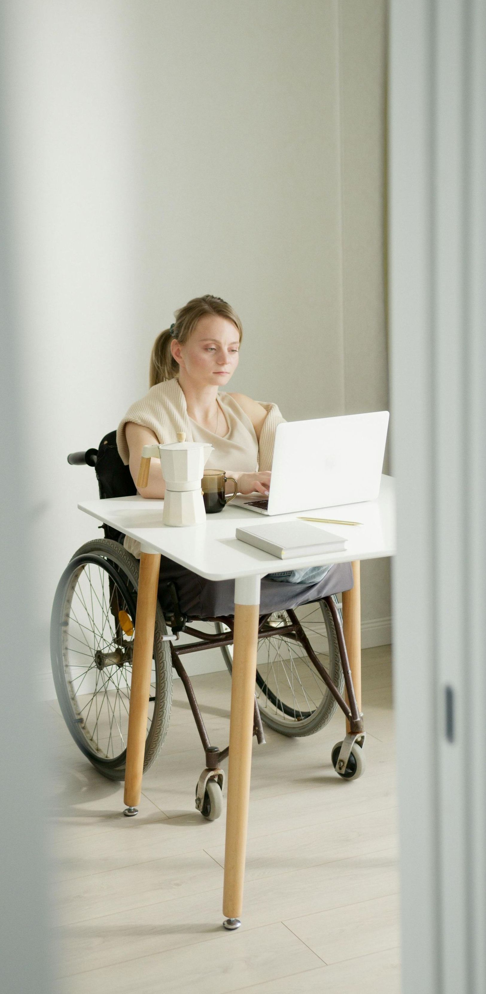 A woman in a wheelchair is sitting at a desk using a laptop computer.