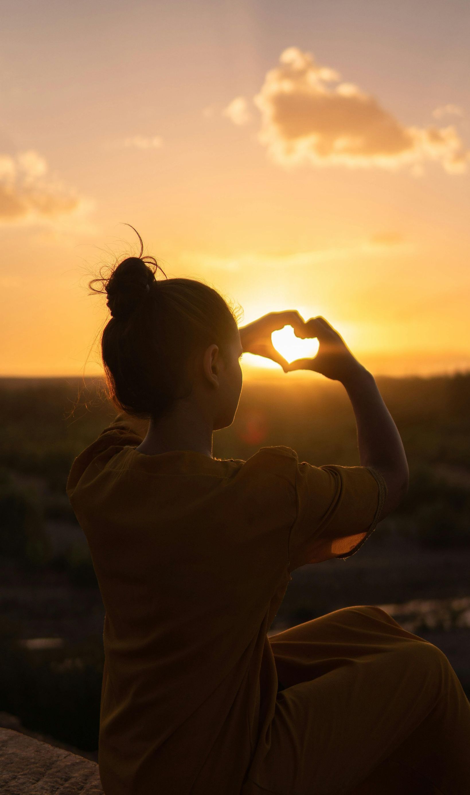 A woman is making a heart shape with her hands at sunset.