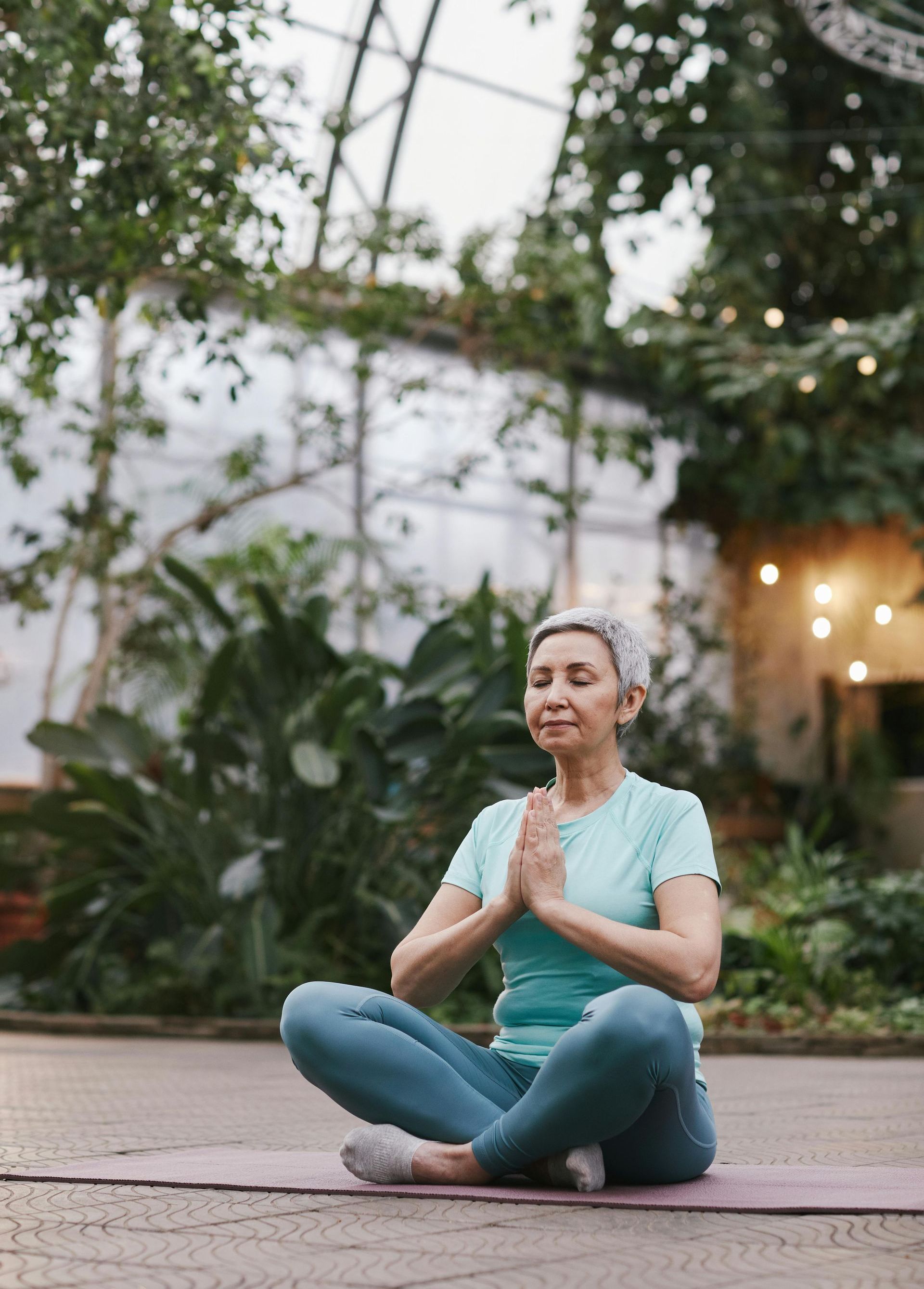 An elderly woman is sitting on a yoga mat in a greenhouse.