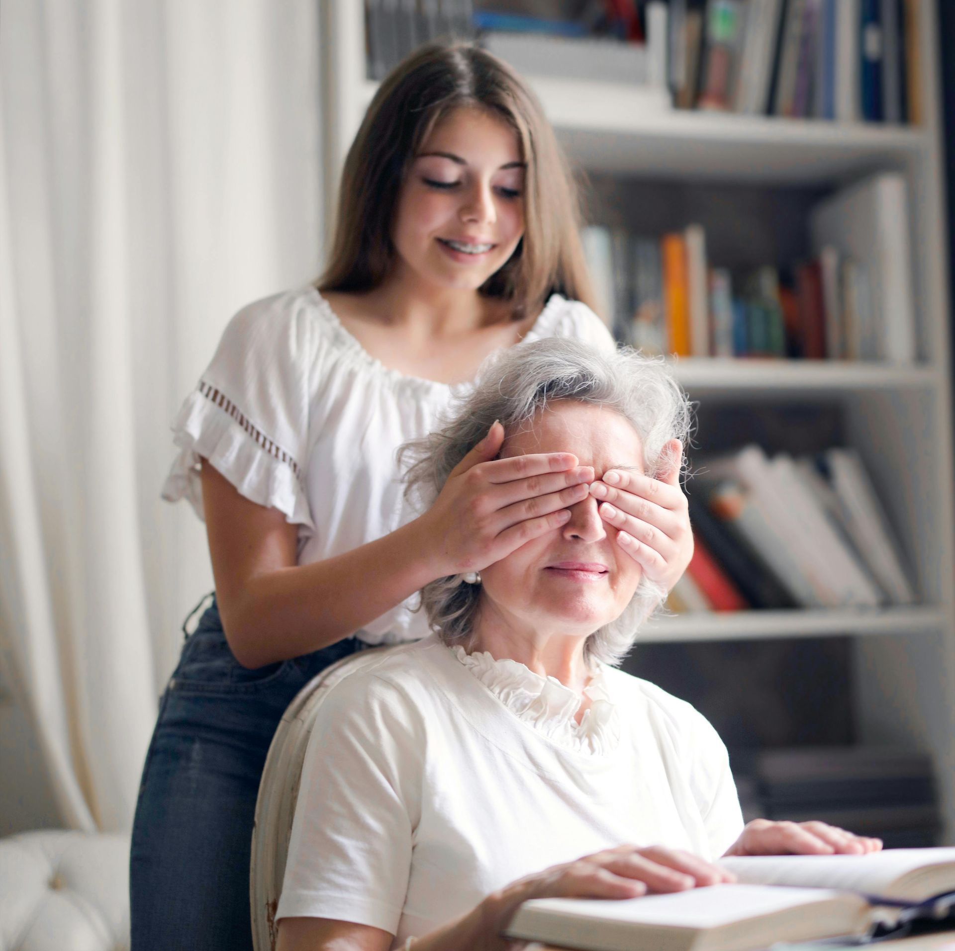 A young woman is covering an older woman 's eyes while she reads a book.