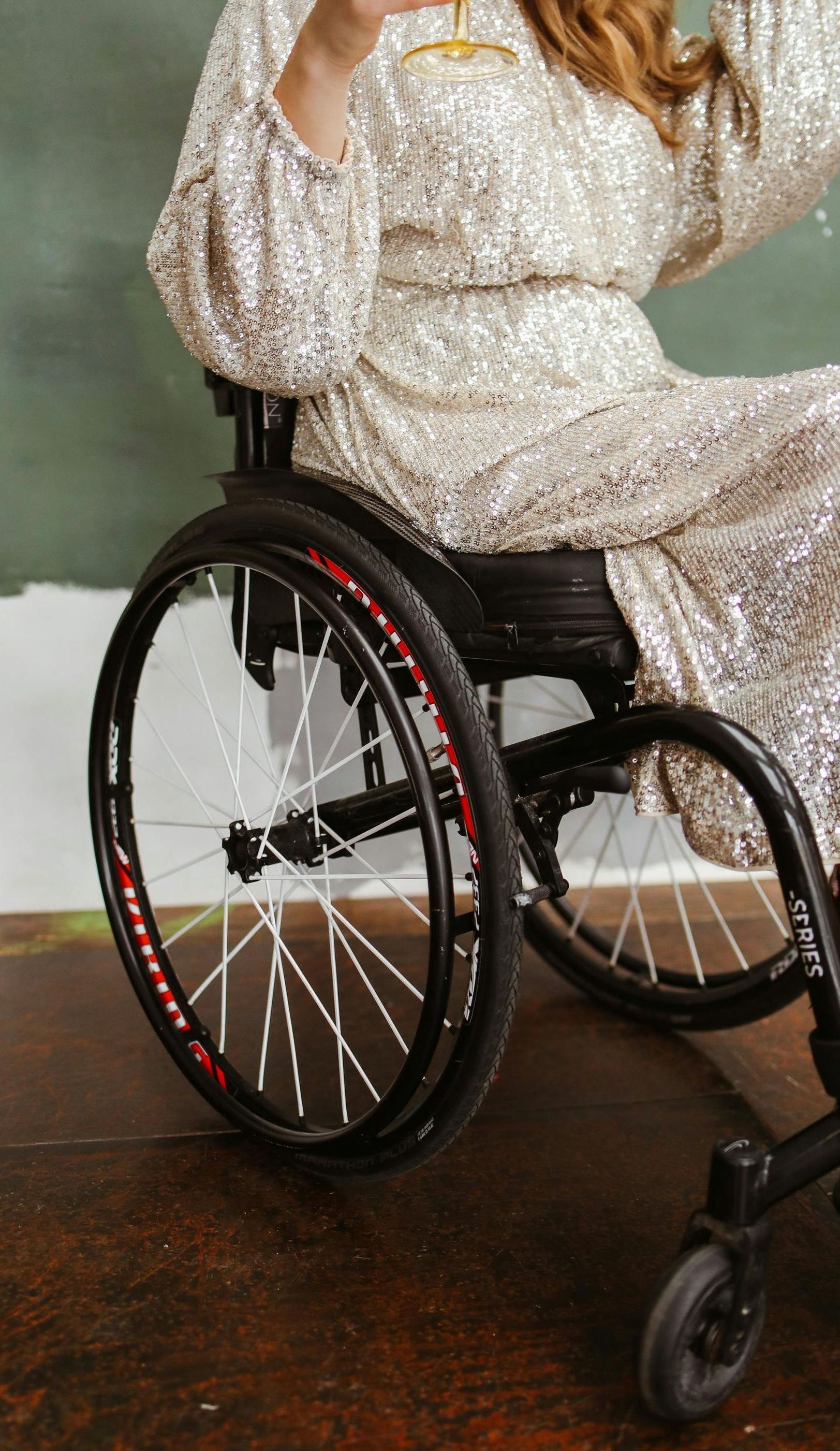 A woman in a sequined dress is sitting in a wheelchair holding a glass of wine.