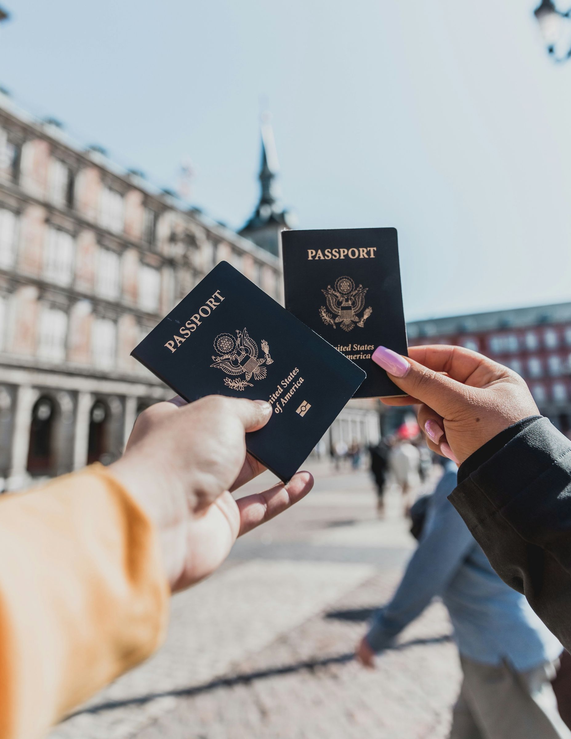 Two people are holding up two passports in front of a building.