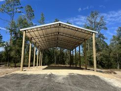 A large metal building is sitting on top of a dirt field in the middle of a forest.