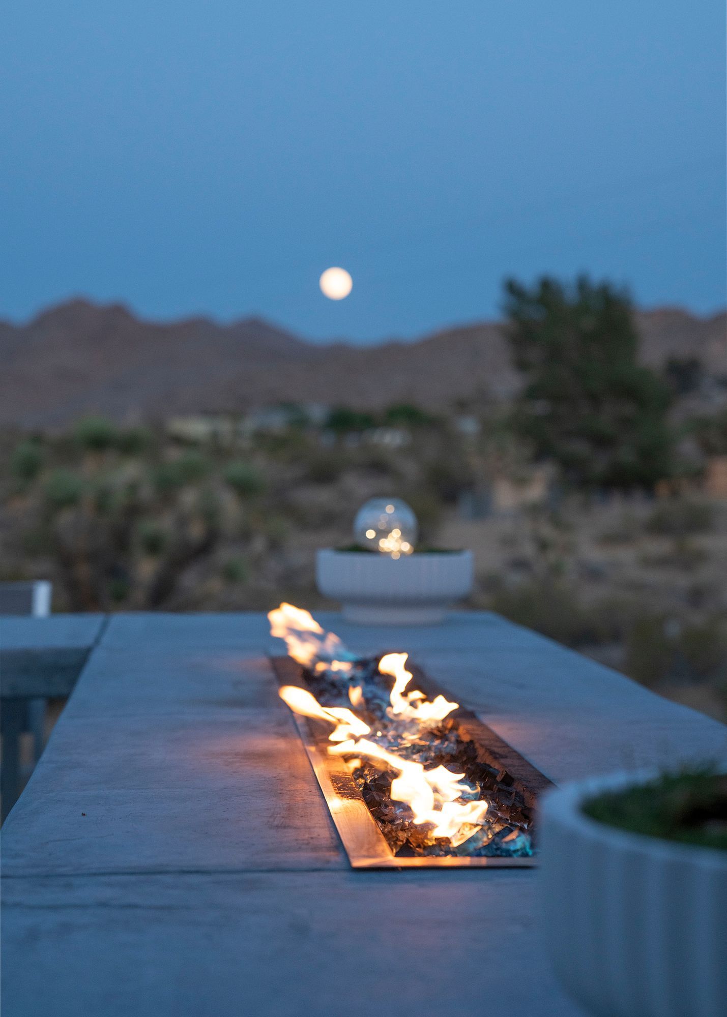 Fire pit ablaze outdoors at dusk, full moon in sky, desert landscape.