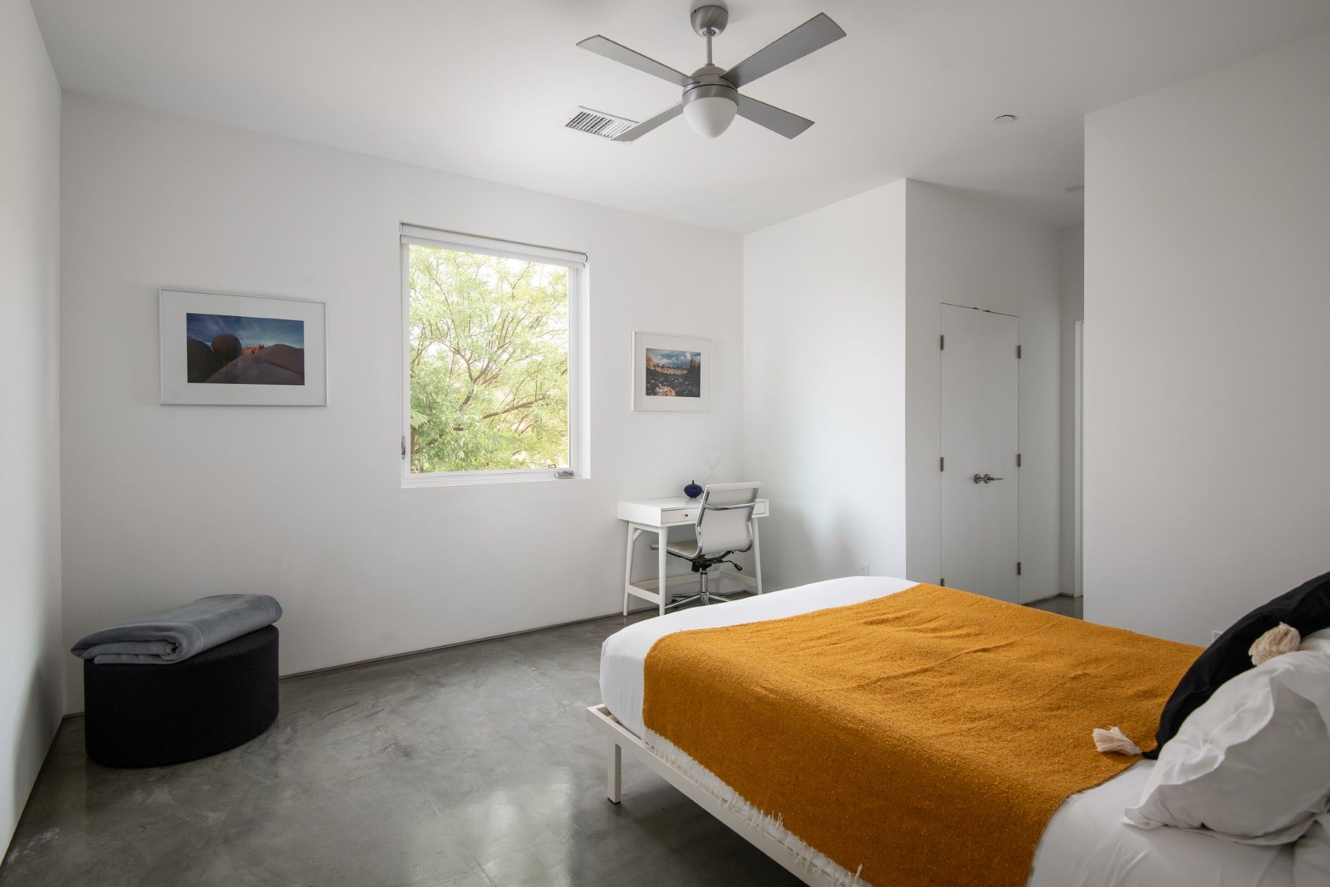 Bedroom with a bed, desk, and window overlooking trees; white walls, concrete floor, and yellow blanket.