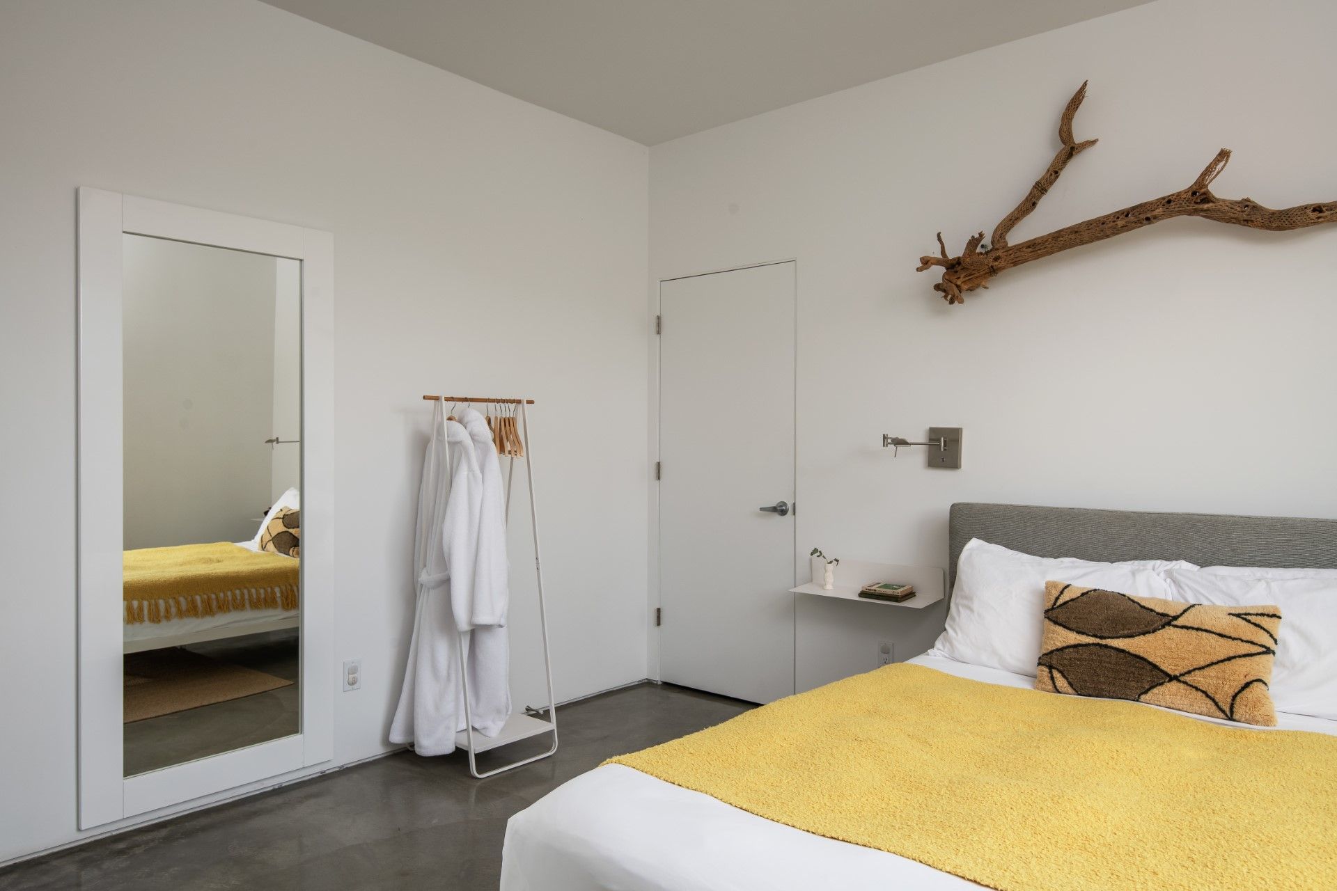 Bedroom with white walls, full-length mirror, bed with yellow cover, driftwood art above the headboard.