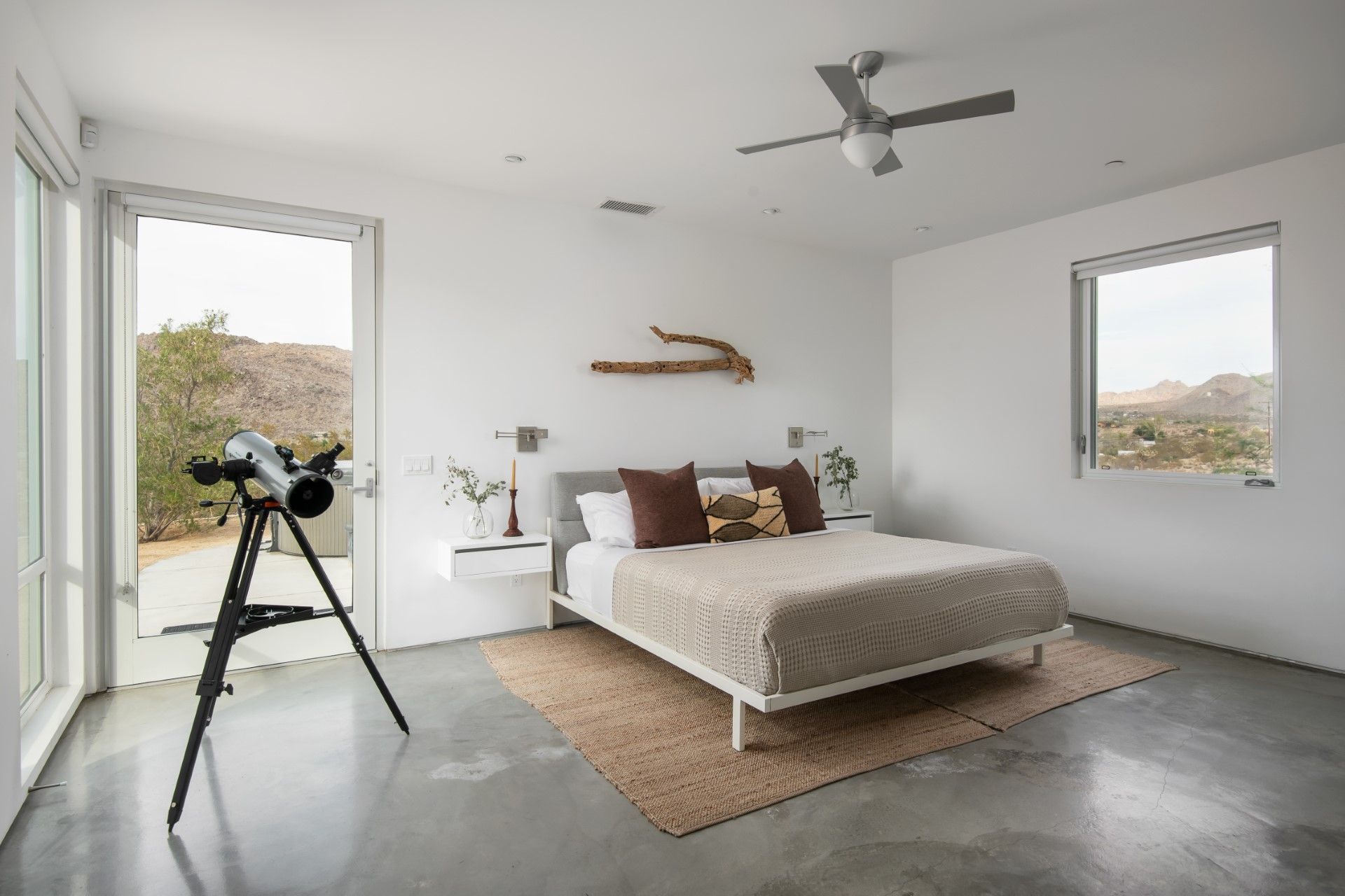 Bedroom with a telescope by a window, bed with brown pillows and blanket, neutral tones.