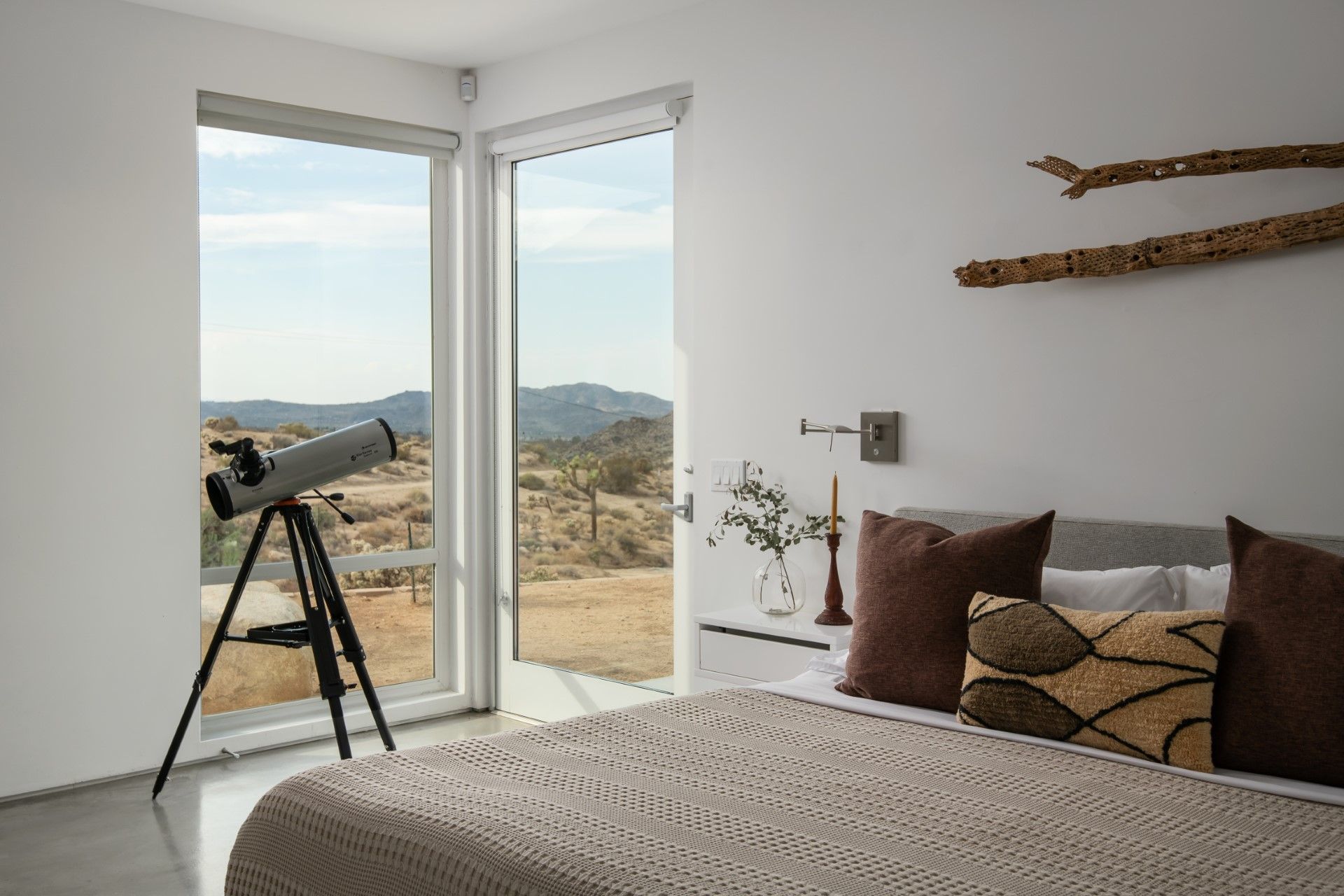 Bedroom with bed, telescope by window overlooking a landscape. White walls, neutral tones, and decorative branches.
