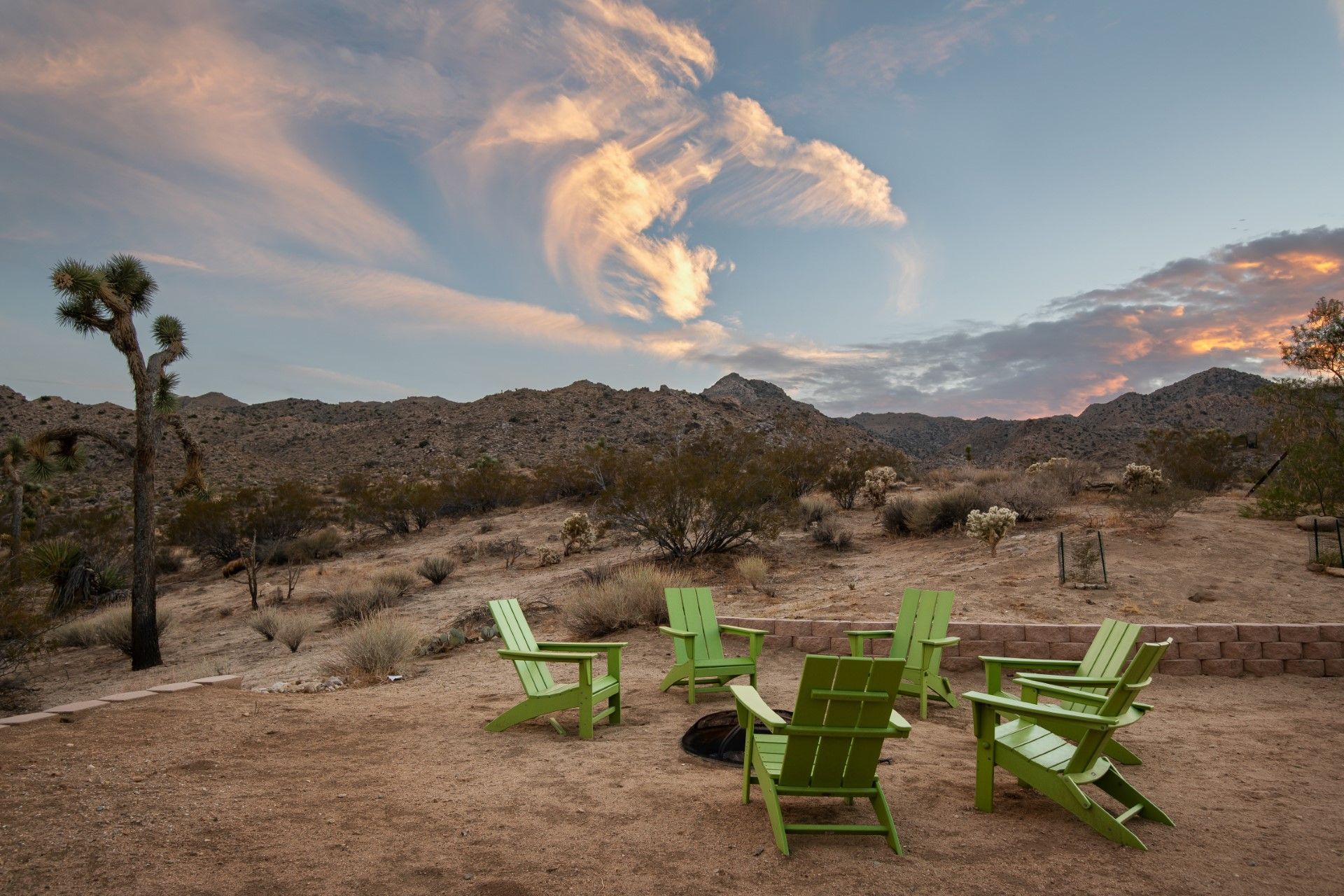Green chairs circle a fire pit in a desert landscape under a colorful sunset sky.