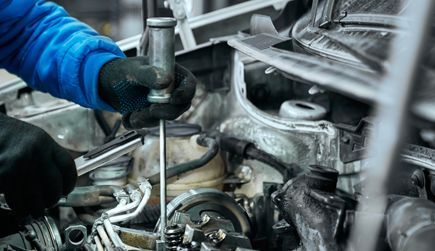 Mechanic working on car engine with a screwdriver, close-up, black gloves, blue sleeves.
