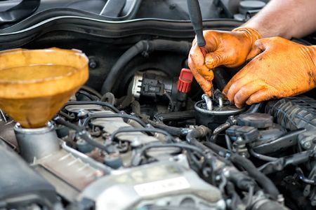 Mechanic with orange gloves working on car engine, funnel visible.